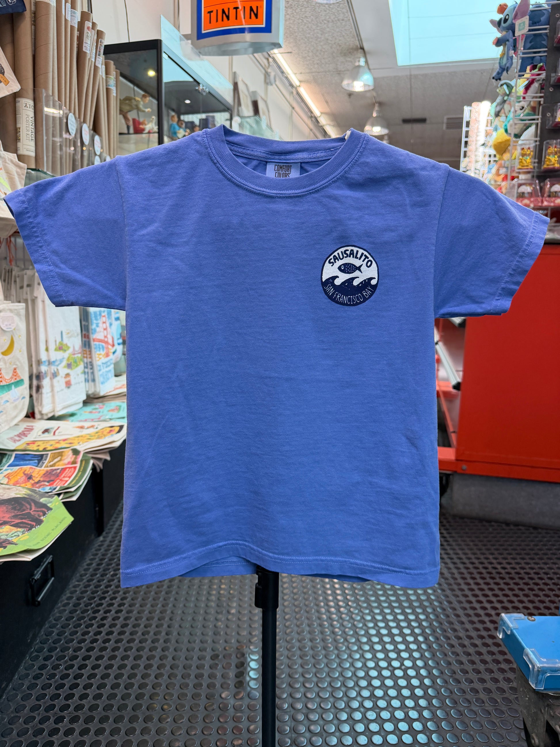 Front of a flo blue short-sleeve kids T-shirt displayed on a stand inside the Sausalito Ferry Co. retail store. The left chest features a small circular white logo with a black design of a fish jumping over ocean waves, surrounded by the text "Sausalito" and "San Francisco Bay."