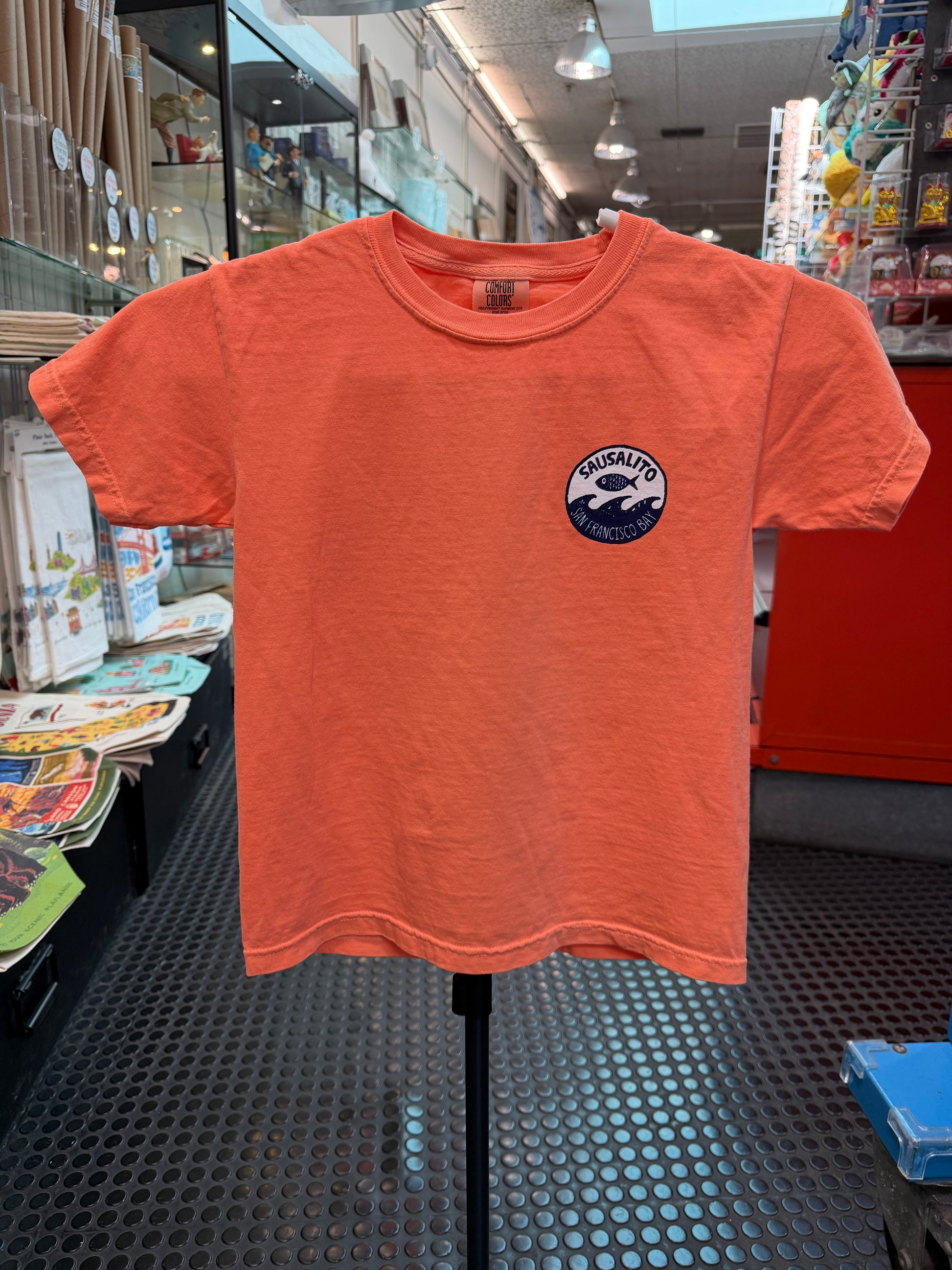 Front of a melon orange short-sleeve kids T-shirt displayed on a stand inside the Sausalito Ferry Co. retail store. The left chest features a small circular navy blue logo with a design of a fish jumping over ocean waves, surrounded by the text "Sausalito" and "San Francisco Bay."