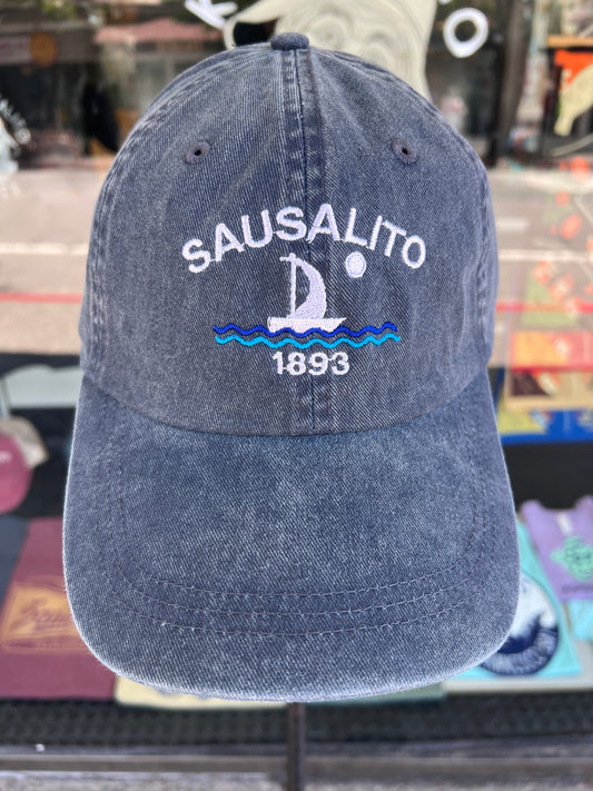 A front view of a washed, pigment-dyed denim blue 100% cotton baseball cap displayed on a stand. The front panel features clean white embroidered block lettering that reads "SAUSALITO" arched over a white sailboat sailing on layered light blue and royal blue stitched waves under a small white full moon, with the year "1893" cleanly embroidered below the waves.