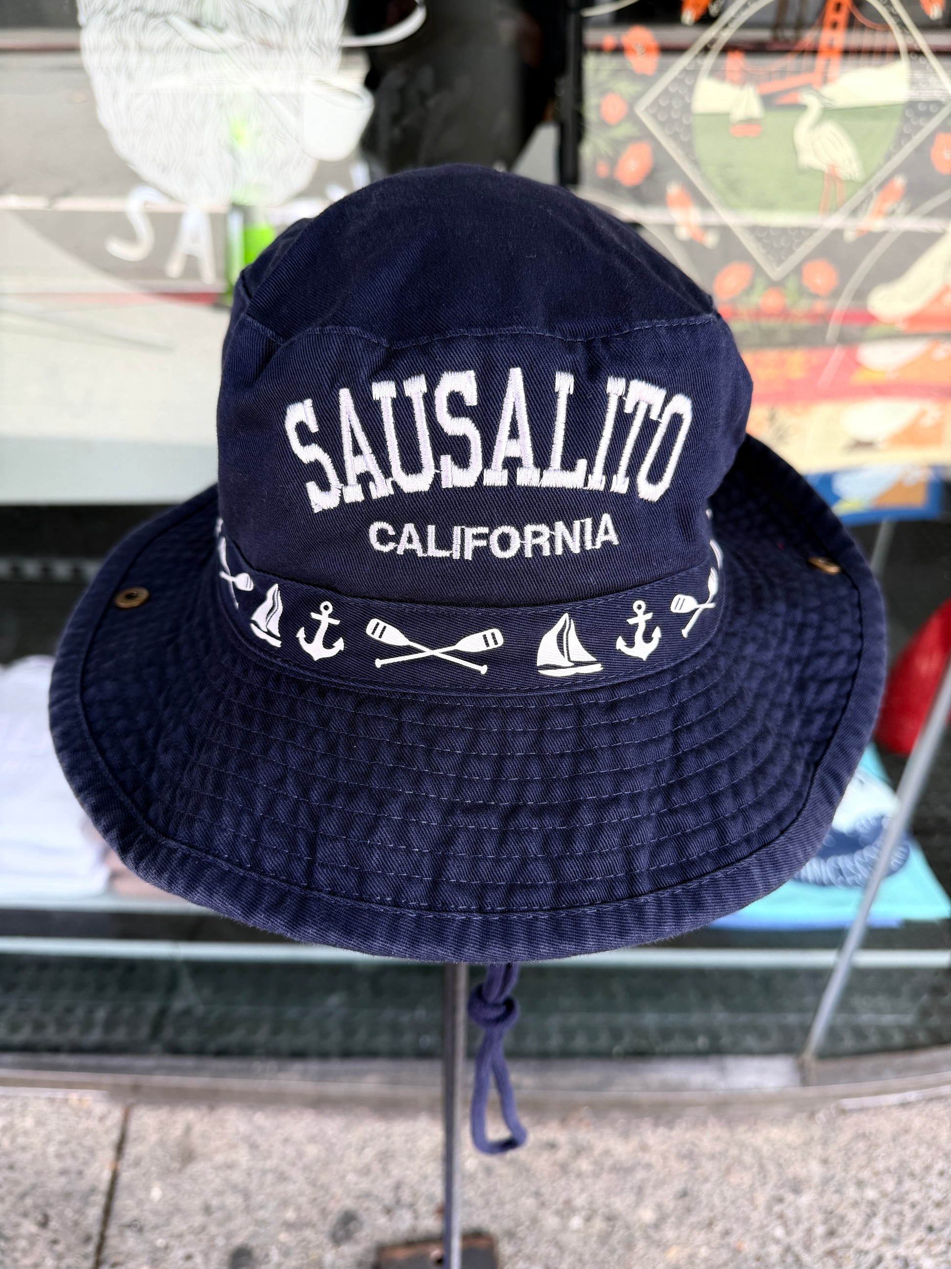 A navy blue cotton bucket hat displayed on a stand. The front features crisp white embroidery reading "SAUSALITO CALIFORNIA" in bold block letters, accented by classic nautical icons including anchors, a sailboat, a ship's wheel, and waves.