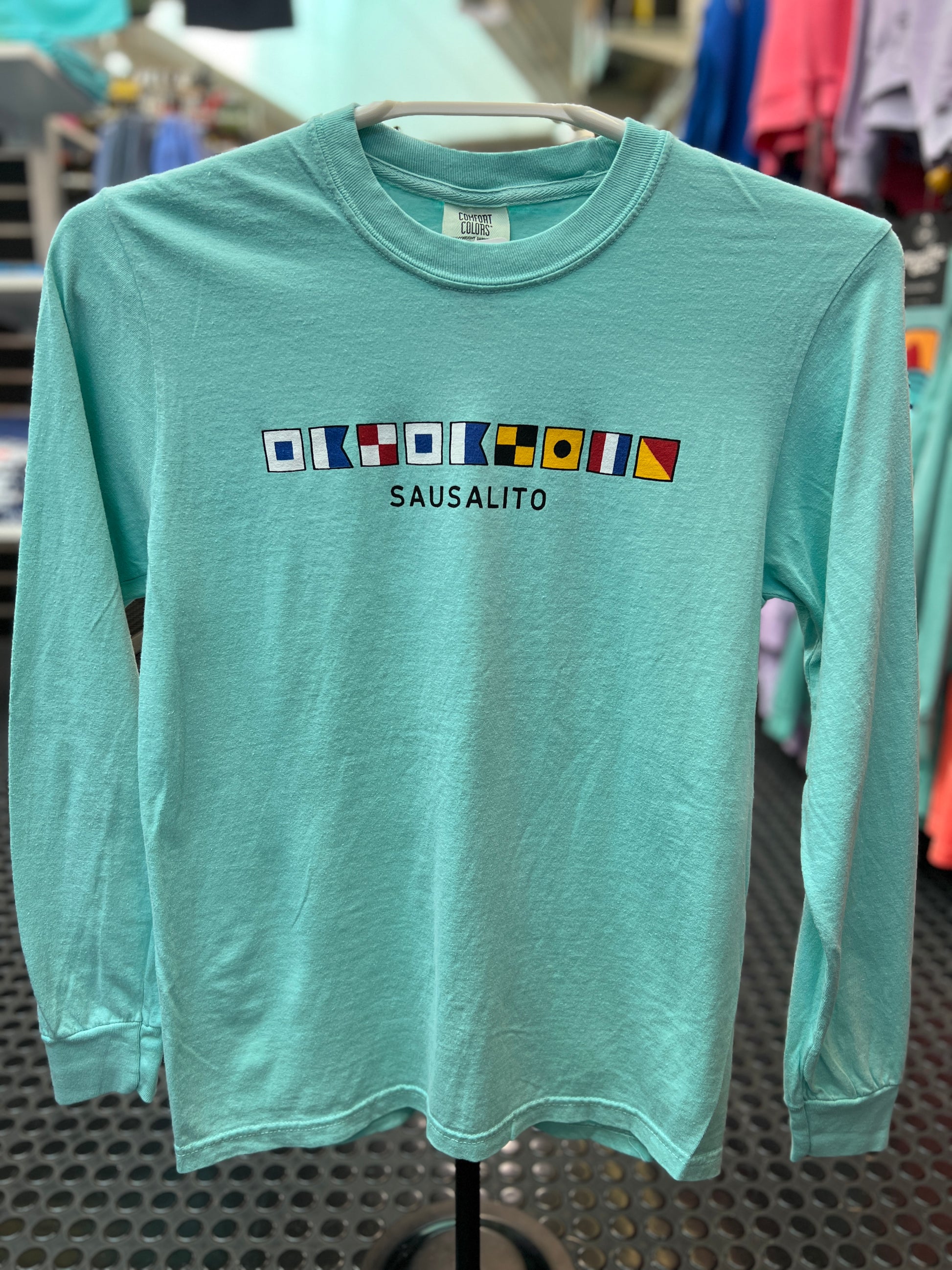 A long sleeve navy blue t-shirt featuring a series of colorful nautical signal flags arranged vertically above the bold white text "SAUSALITO." The nautical flags spell out "Sausalito".