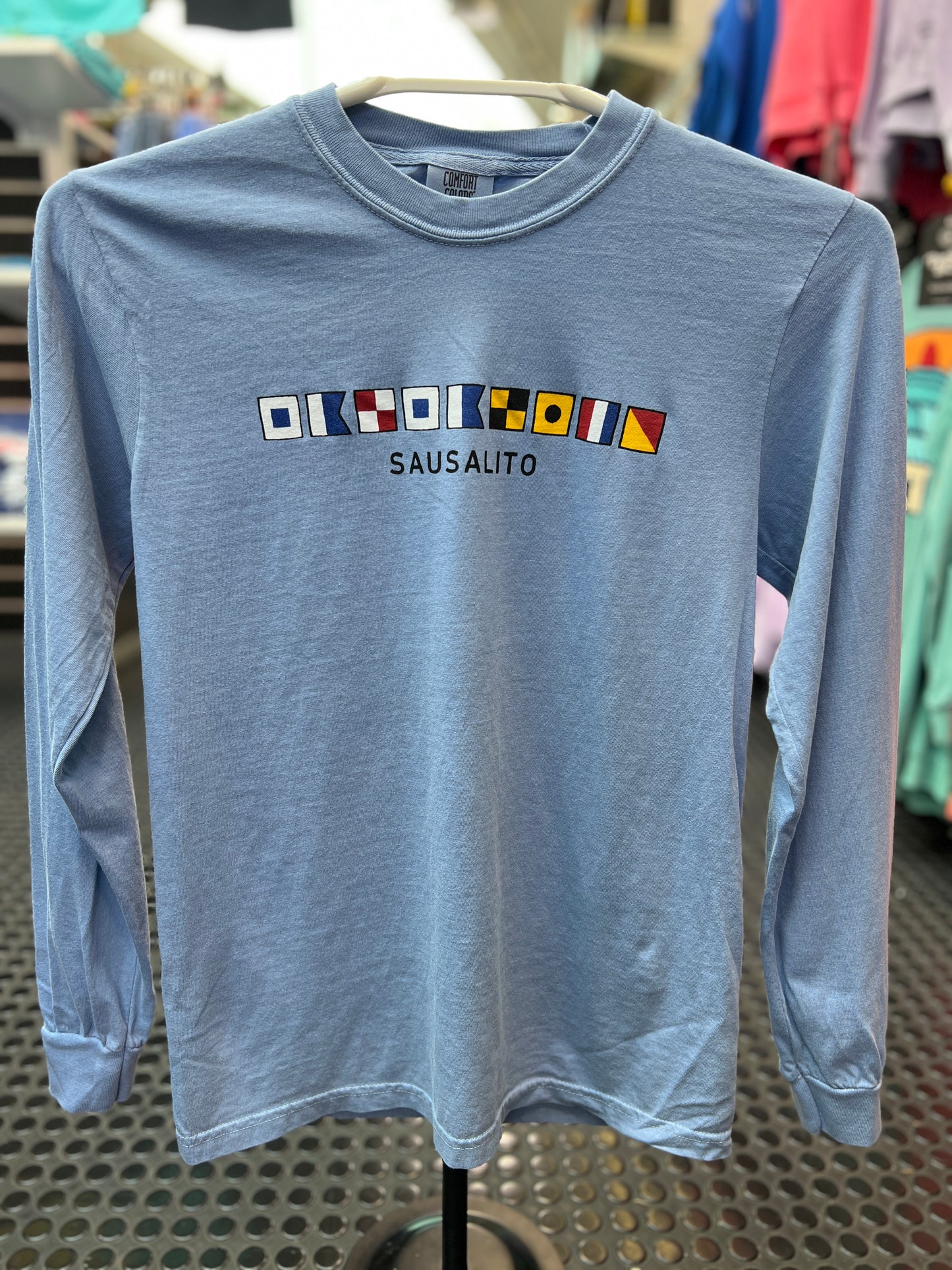 A Comfort Colors short sleeve t-shirt featuring a horizontal row of colorful maritime nautical signal flags that spell out 'Sausalito' across the chest."