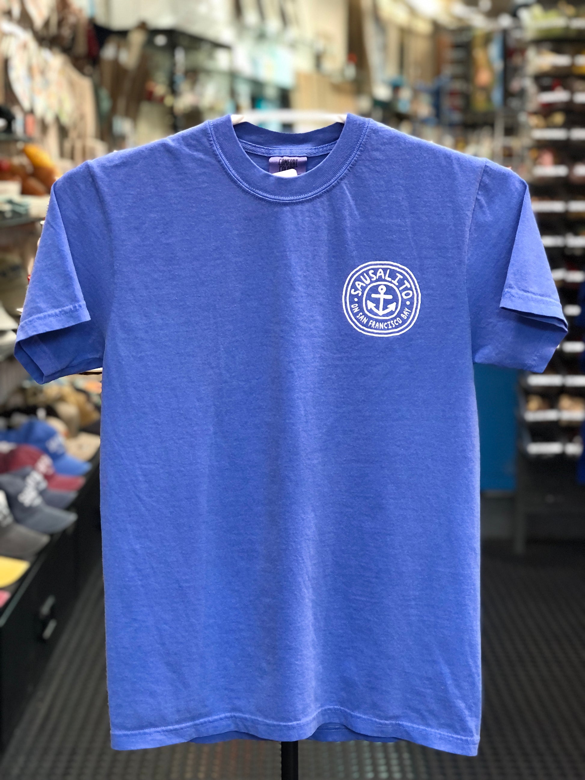 Blue Comfort Colors t-shirt with a circular pocket logo featuring an anchor and text on a hanger in a store. 