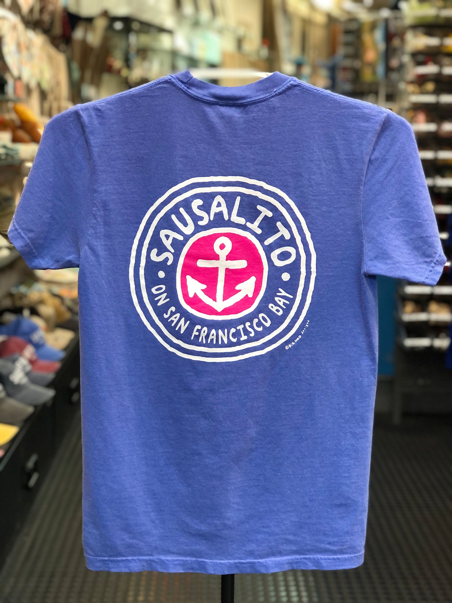 Blue Comfort Colors t-shirt with a circular logo featuring an anchor and text on a hanger in a store. 