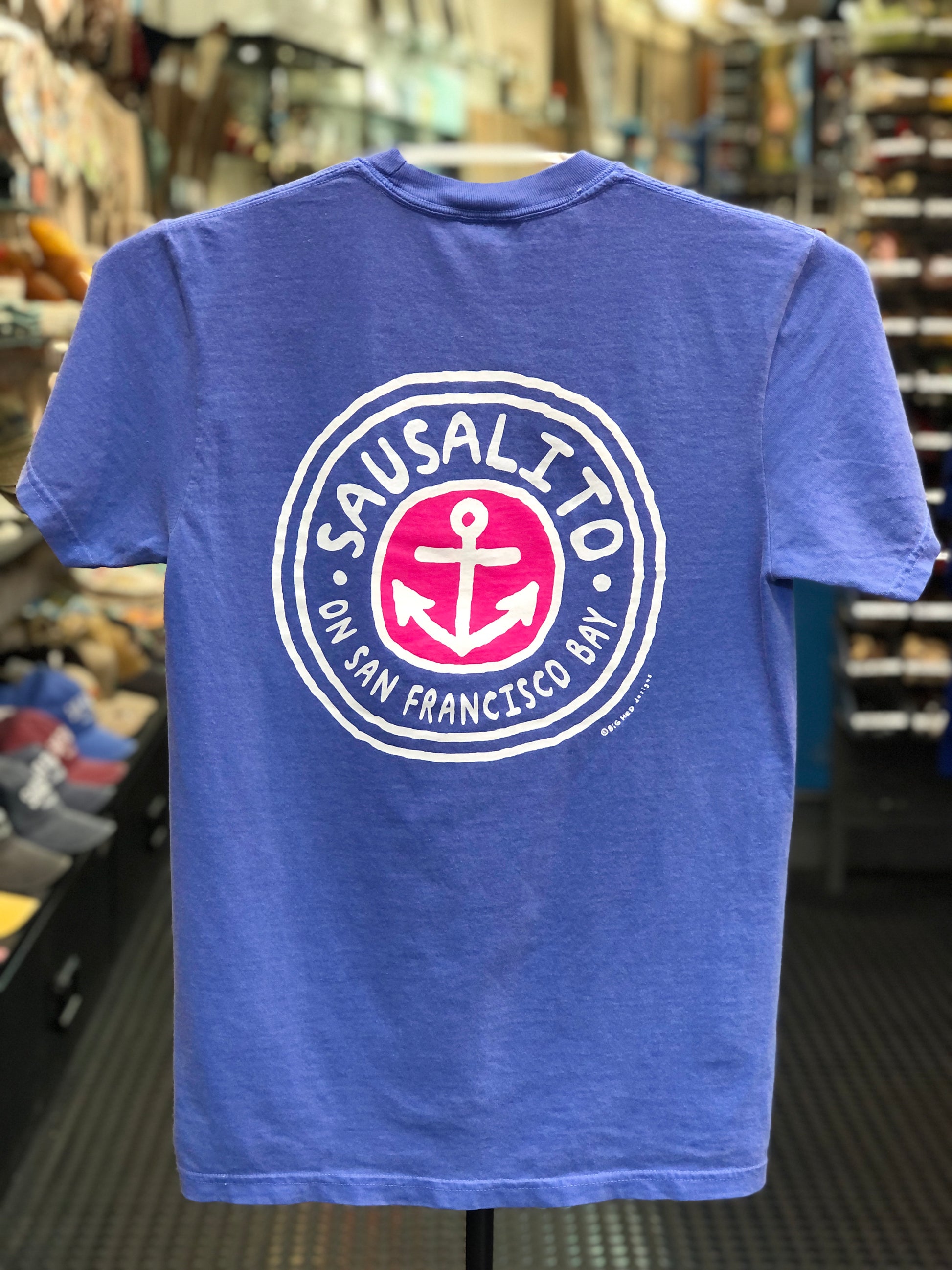 Blue Comfort Colors t-shirt with a circular logo featuring an anchor and text on a hanger in a store. 