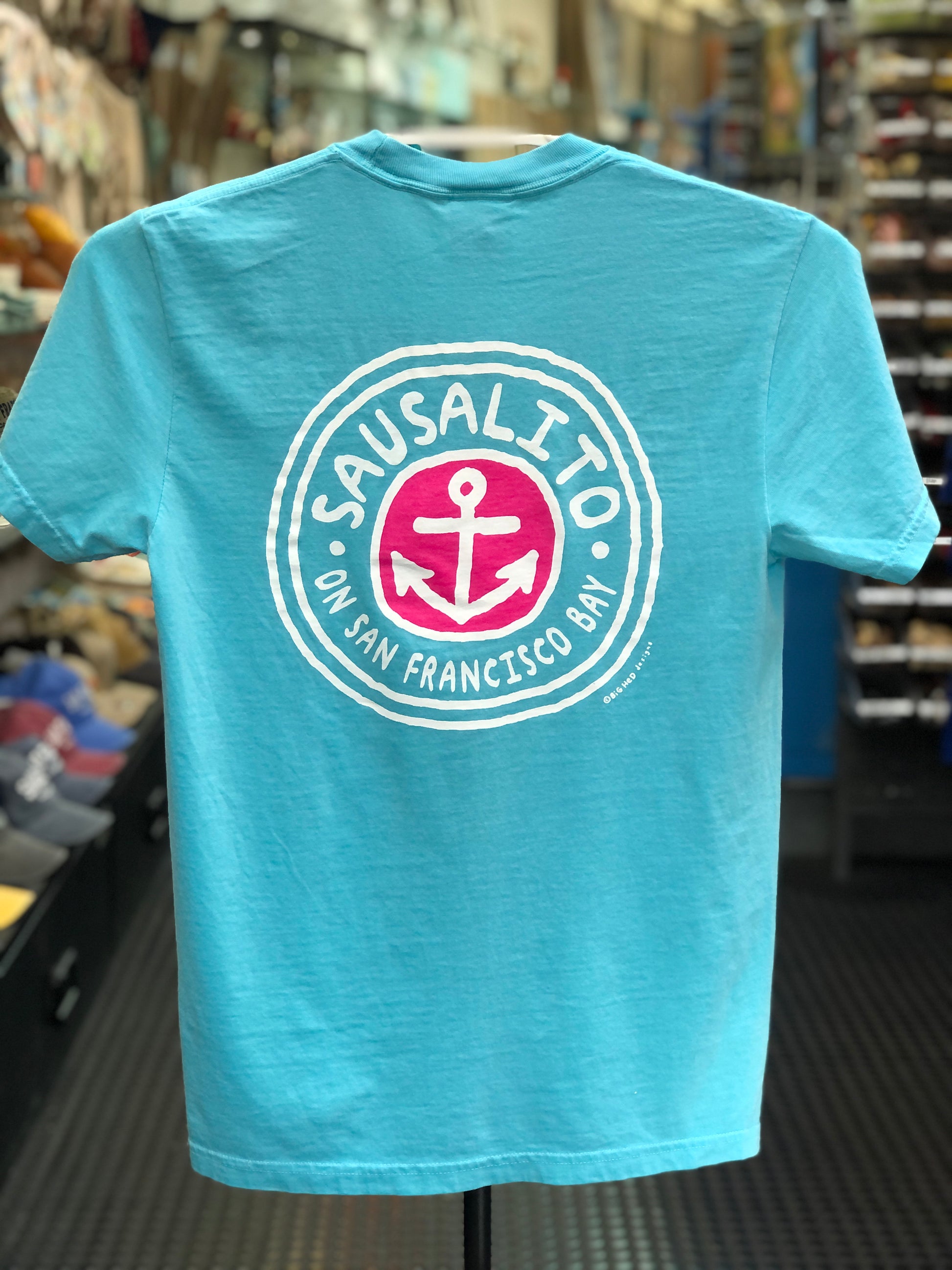 Lagoon blue Comfort Colors t-shirt featuring a circular "Sausalito On San Francisco Bay" anchor logo printed on the back, displayed on a clothing hanger.