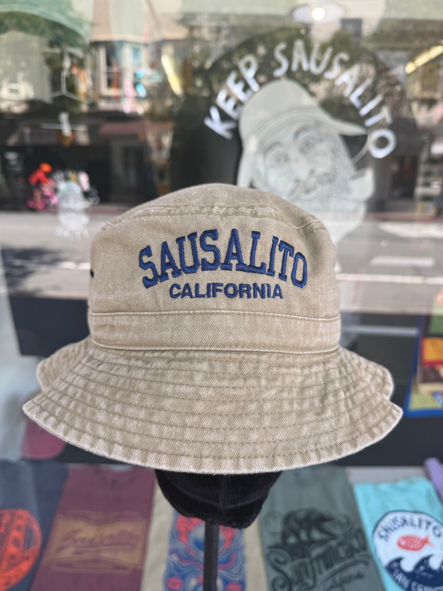 A front view of a beige, over-washed 100% cotton bucket hat displayed on a black mannequin stand in front of a store window. The front features the words "SAUSALITO" cleanly embroidered in blue block letters arched over "CALIFORNIA" in smaller blue text. The reflection in the window shows a vintage style illustration under the arched words "KEEP SAUSALITO SALTY".