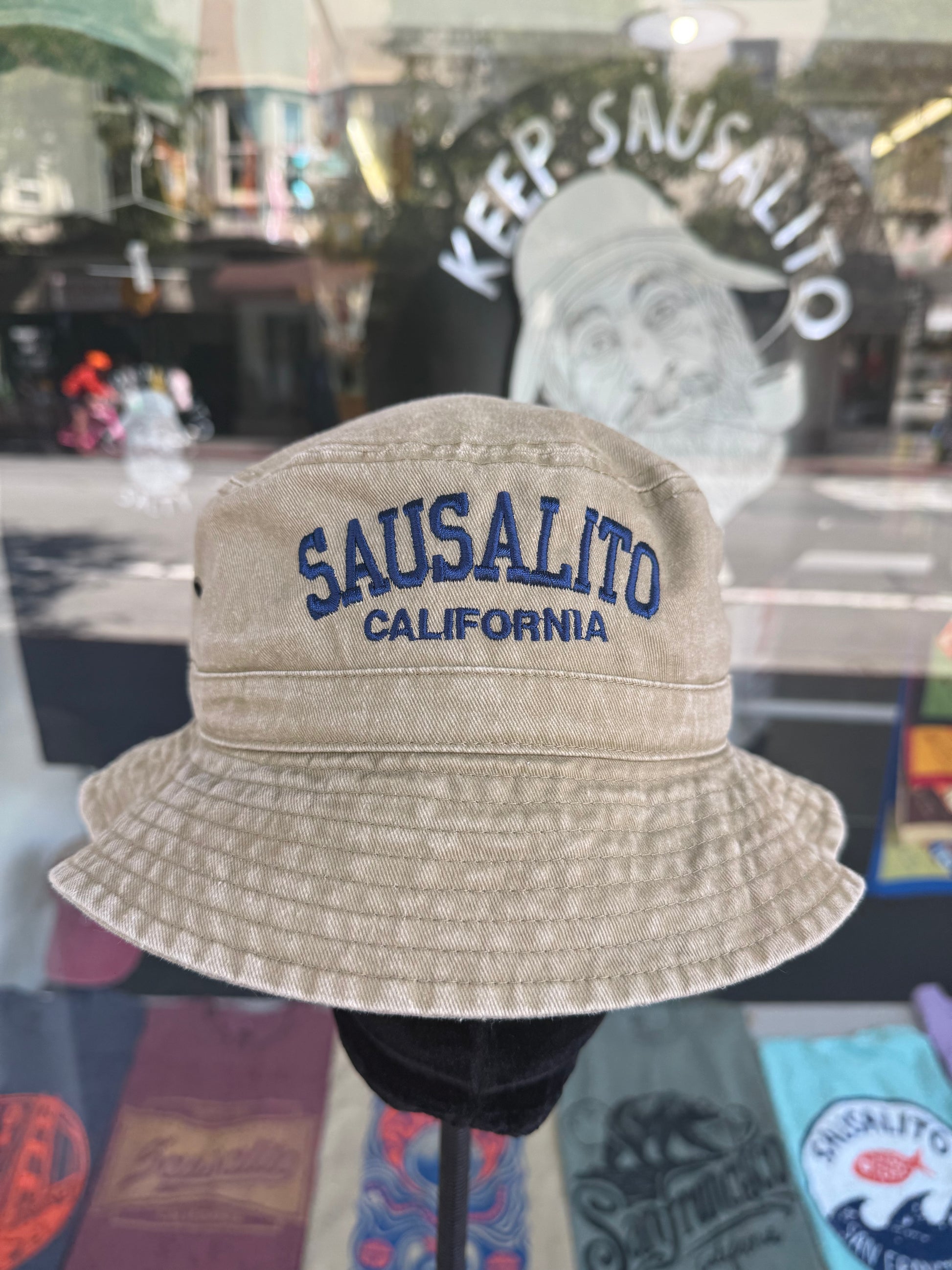A front view of a beige, over-washed 100% cotton bucket hat displayed on a black mannequin stand in front of a store window. The front features the words "SAUSALITO" cleanly embroidered in blue block letters arched over "CALIFORNIA" in smaller blue text. The reflection in the window shows a vintage style illustration under the arched words "KEEP SAUSALITO SALTY".