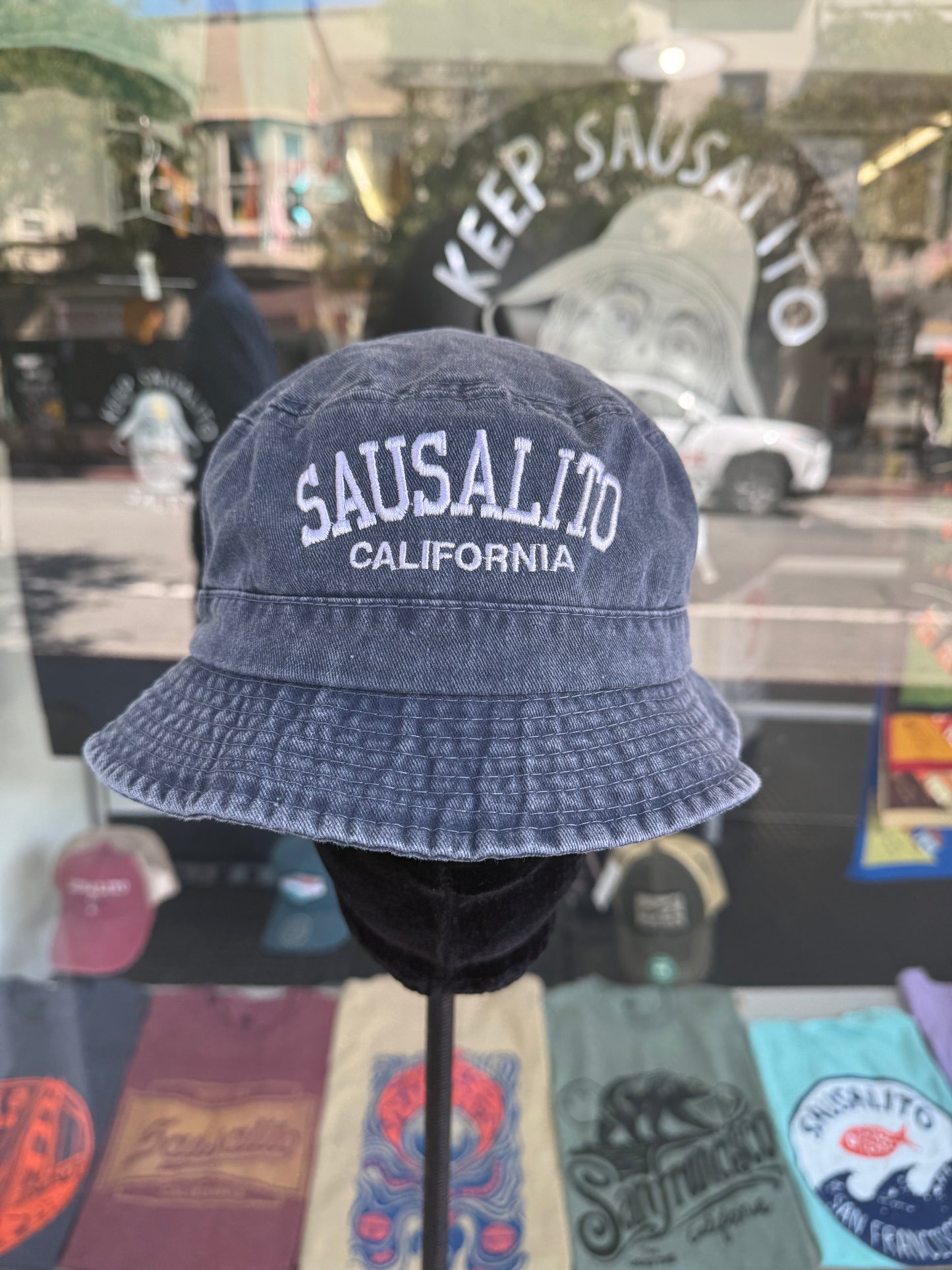 A front view of a navy blue, over-washed 100% cotton bucket hat displayed on a black mannequin stand in front of a store window. The front features the words "SAUSALITO" cleanly embroidered in white block letters arched over "CALIFORNIA" in smaller white text. In the background below the hat, a colorful row of folded graphic T-shirts is visible, while the window reflection shows the storefront sign "KEEP SAUSALITO SALTY".