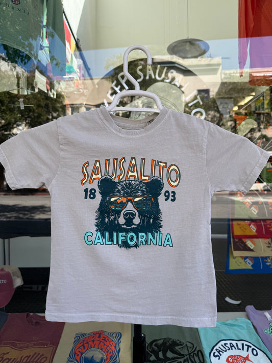 White t-shirt with bear graphic and "Sausalito 1893 California" text on a hanger in front of a store window.