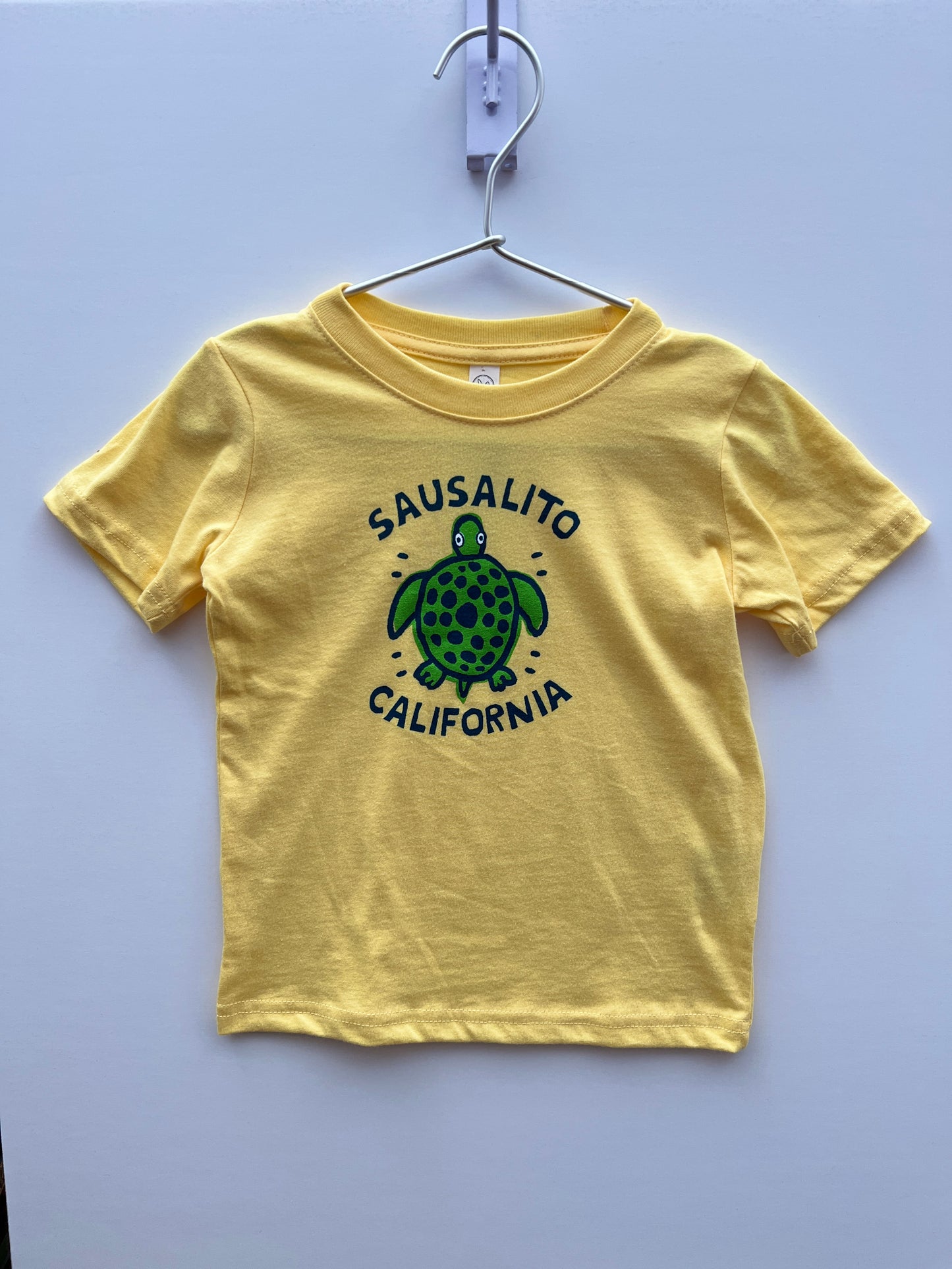 Yellow kids' t-shirt with green turtle design and "Sausalito California" text, hanging on white background