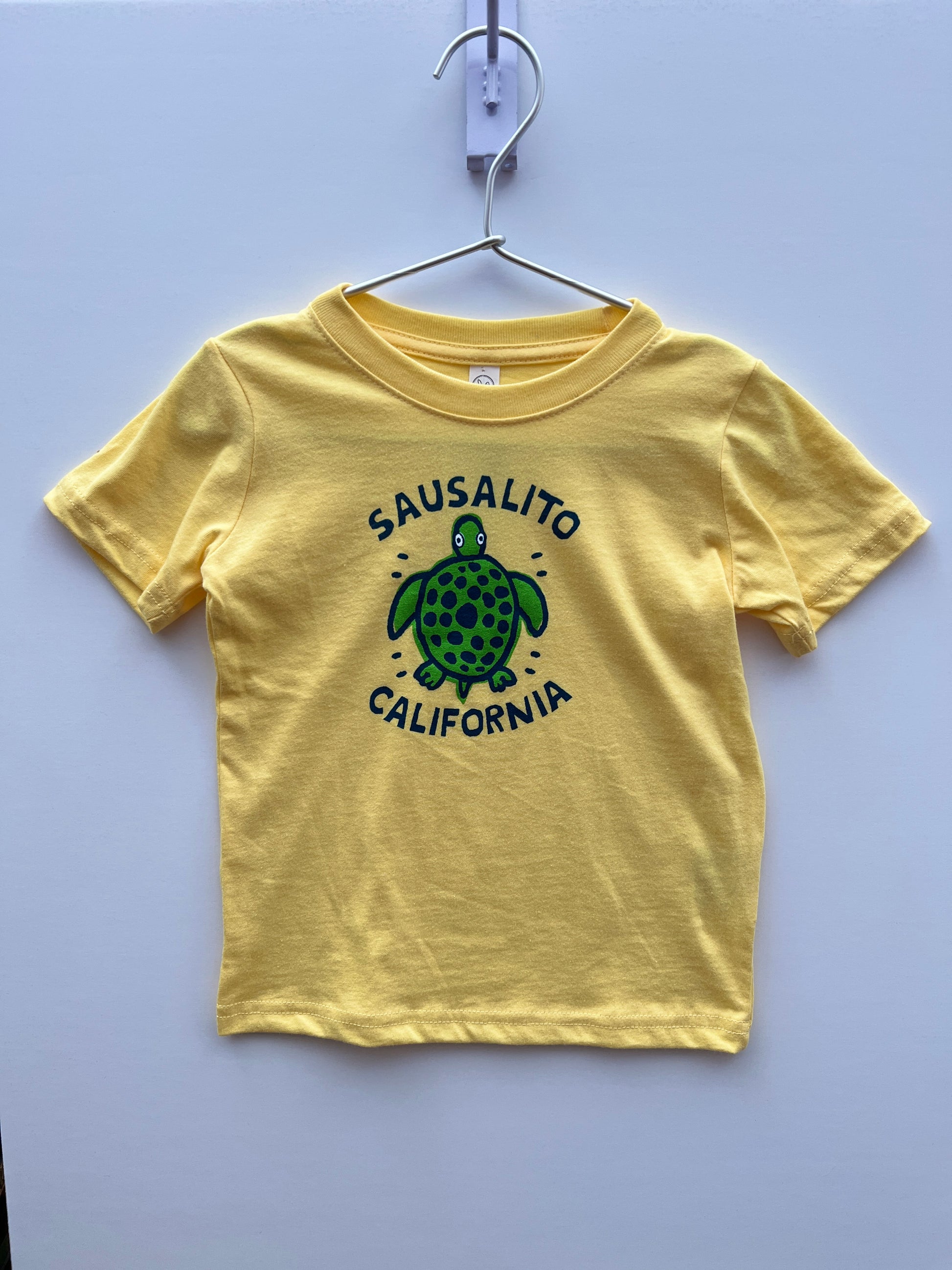 Yellow kids' t-shirt with green turtle design and "Sausalito California" text, hanging on white background