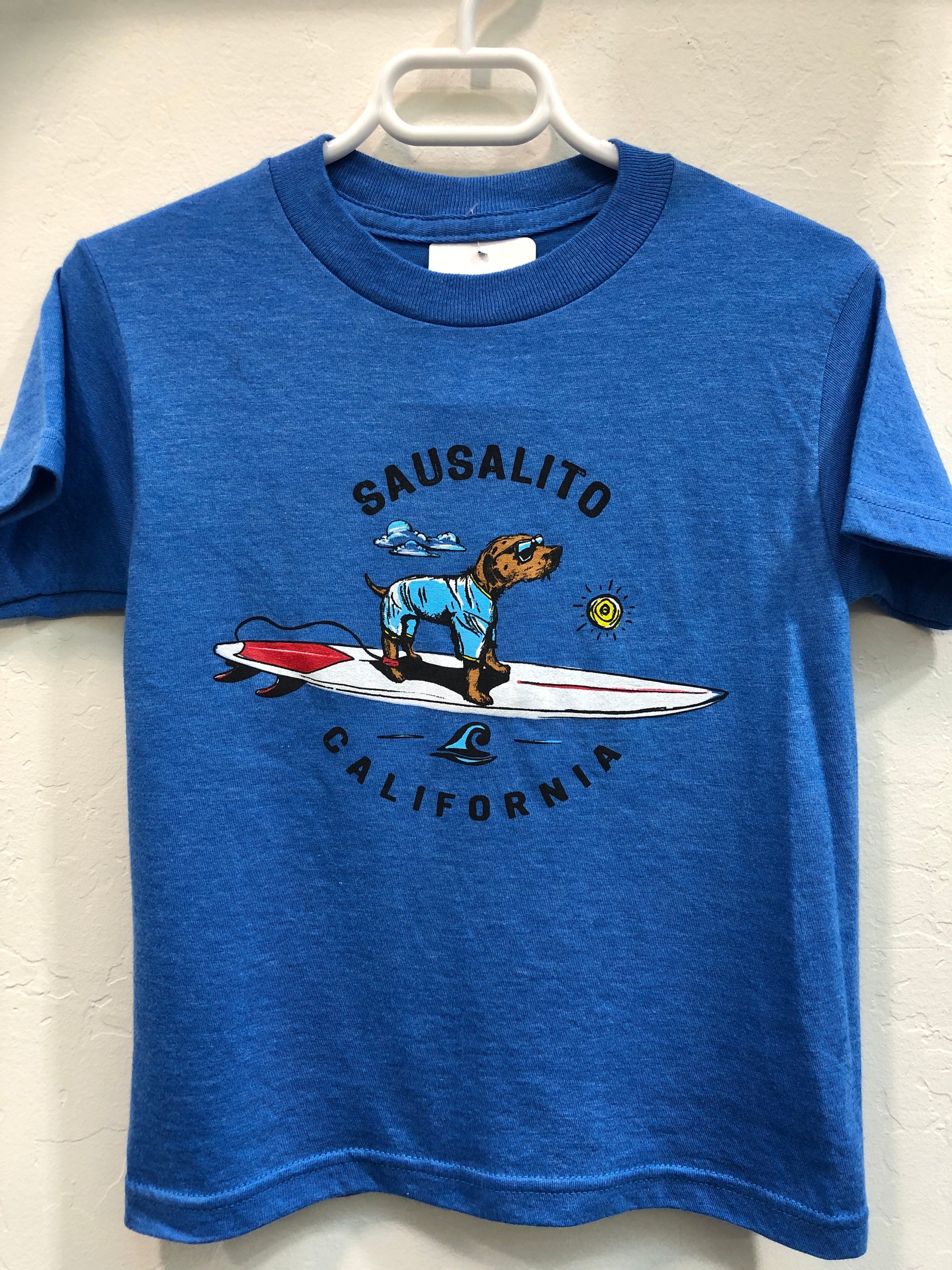 Blue kids' t-shirt with cartoon surfing dog graphic, "Sausalito California" text, hanging on white hanger