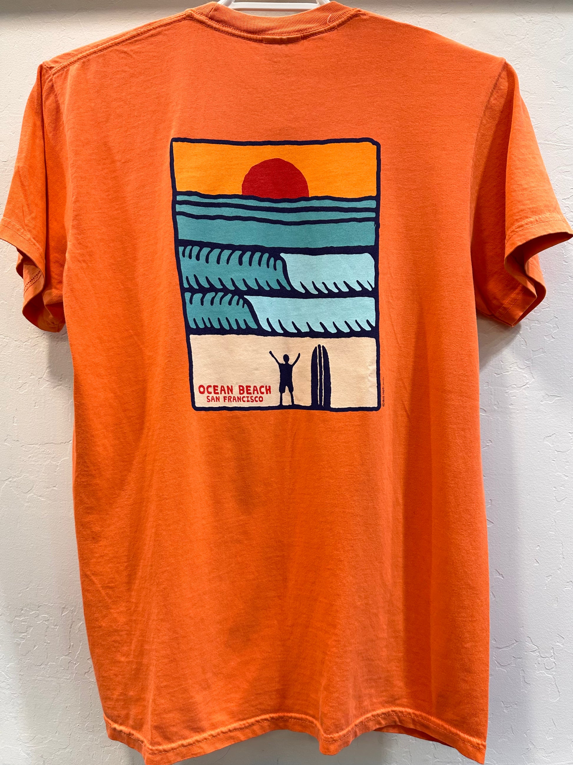 The back of a terracotta orange t-shirt featuring a large rectangular graphic. The design displays a dark red setting sun, vibrant blue and teal ocean waves, and the black silhouette of a surfer standing with arms raised next to their surfboard on the shore, captioned "Ocean Beach San Francisco."