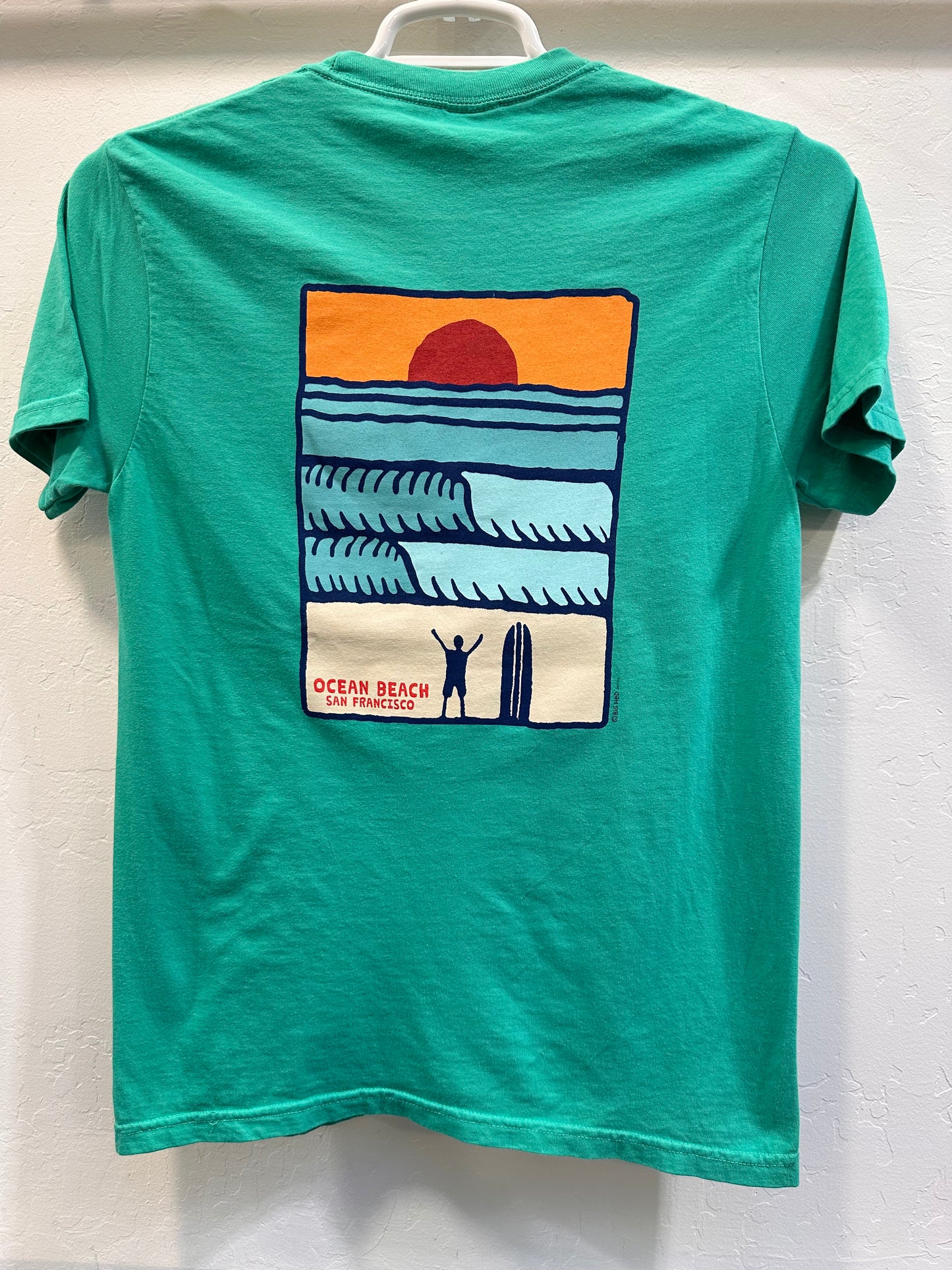 The back of a vibrant green t-shirt showing a rectangular retro graphic of a dark red setting sun over blue ocean waves. A silhouette of a surfer stands on the sandy shore next to a surfboard with arms raised in celebration, captioned "Ocean Beach San Francisco."