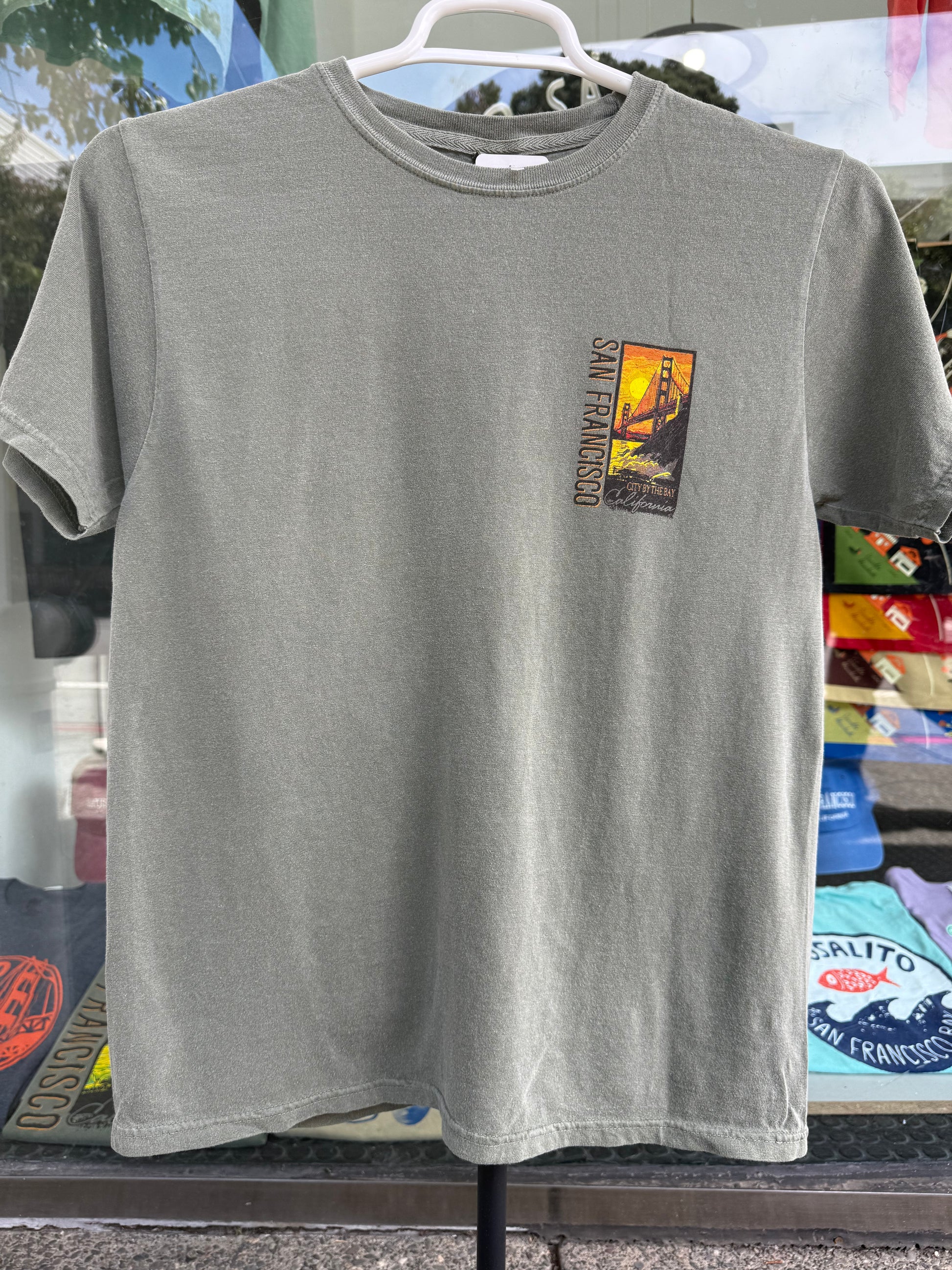 Front view of a sage green garment-dyed t-shirt on a hanger, featuring a small, vertical rectangular chest graphic on the left side with a vibrant orange sunset silhouette of the Golden Gate Bridge and the text "SAN FRANCISCO".