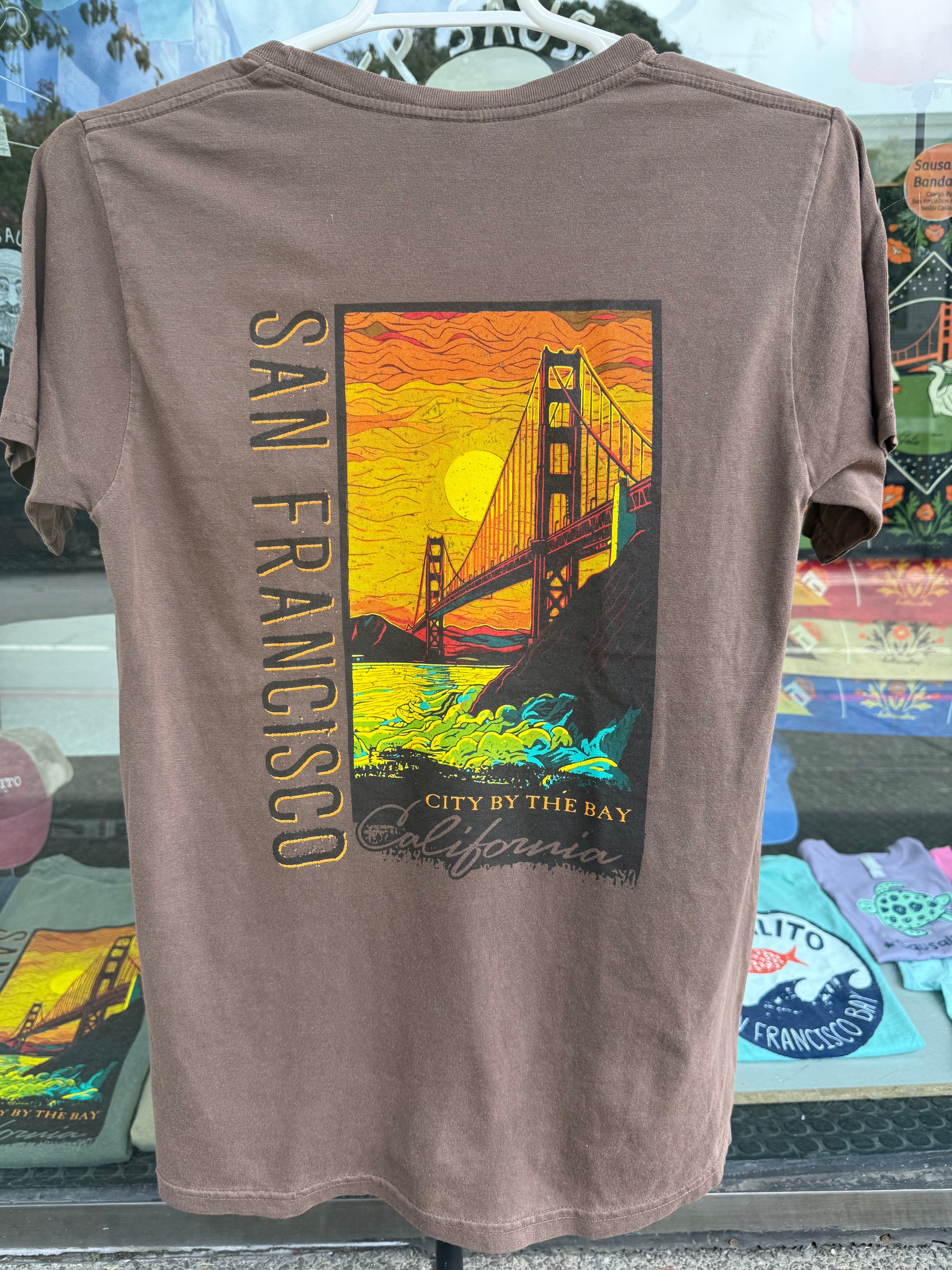 Back view of a coffee bean brown garment-dyed t-shirt featuring a large, vibrant rectangular graphic of the Golden Gate Bridge against a brilliant orange and yellow sunset sky with crashing ocean waves below, alongside bold vertical text that reads "SAN FRANCISCO".