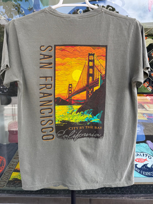Back view of a sage green garment-dyed t-shirt featuring a large, vibrant rectangular graphic of the Golden Gate Bridge against a brilliant orange and yellow sunset sky with crashing ocean waves below, alongside bold vertical text that reads "SAN FRANCISCO".