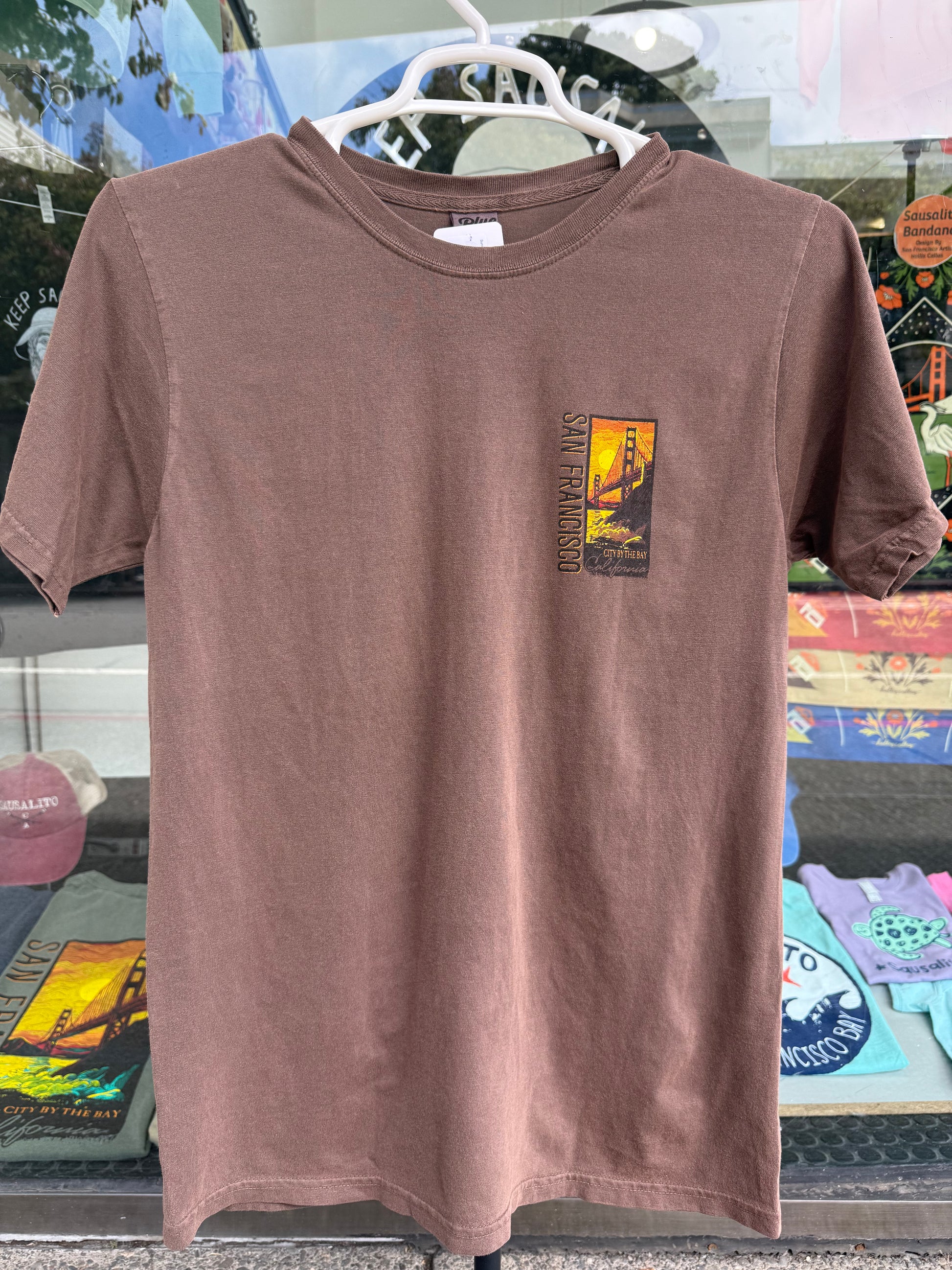 Front view of a coffee bean brown garment-dyed t-shirt on a hanger, featuring a small, vertical rectangular chest graphic on the left side with a vibrant orange sunset silhouette of the Golden Gate Bridge and the text "SAN FRANCISCO".