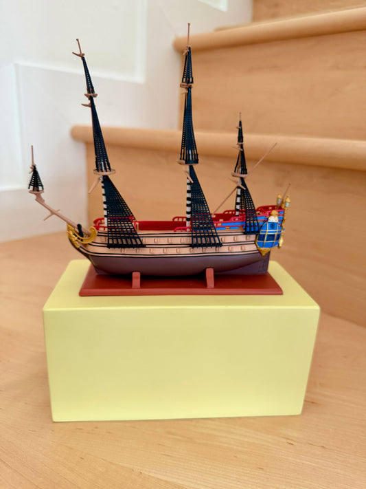 Detailed collectible model of The Unicorn sailing ship from The Adventures of Tintin, displayed on a wooden stand with three masts and blue and red hull accents.