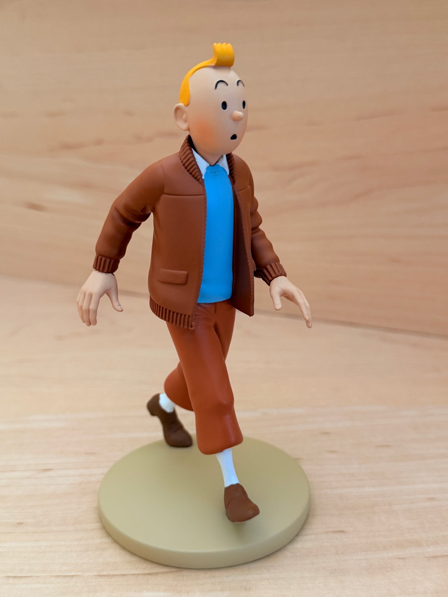 A 12 cm hand-painted resin figure of Tintin from the comic The Castafiore Emerald. He is mid-stride on a round tan base, wearing a brown zippered aviator jacket with a dark brown collar, a bright blue sweater underneath, brown trousers, and white socks with brown shoes.