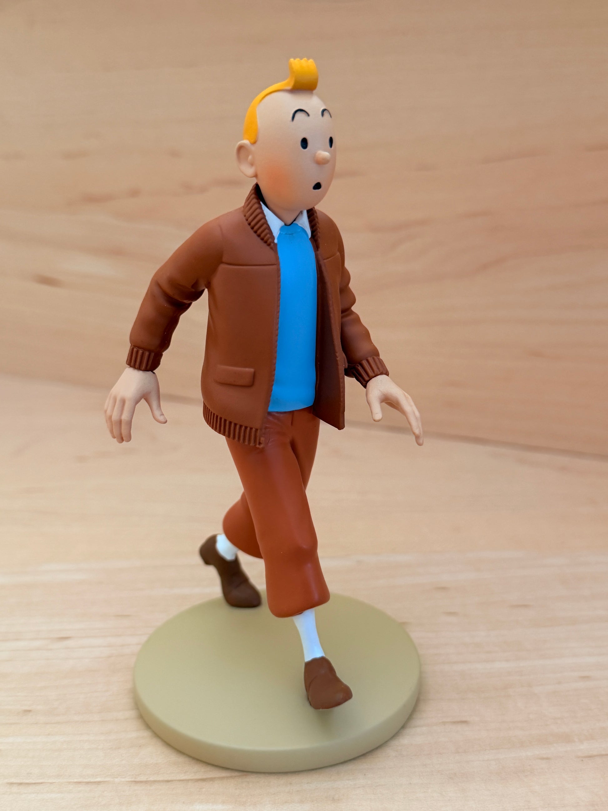 A 12 cm hand-painted resin figure of Tintin from the comic The Castafiore Emerald. He is mid-stride on a round tan base, wearing a brown zippered aviator jacket with a dark brown collar, a bright blue sweater underneath, brown trousers, and white socks with brown shoes.