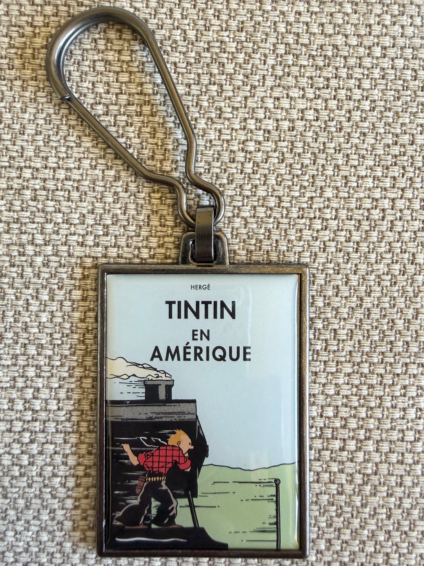 A front close-up view of an officially authorized, rectangular metal Tintin keyring resting on a textured beige fabric surface. The keyring features a durable, scratch-resistant resin-coated front cover from "Tintin in America," showing Tintin dressed in a red plaid shirt, balanced precariously on the side of a speeding steam locomotive as he peers forward.