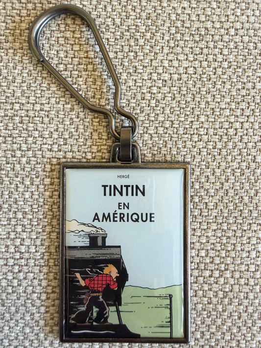 A front close-up view of an officially authorized, rectangular metal Tintin keyring resting on a textured beige fabric surface. The keyring features a durable, scratch-resistant resin-coated front cover from "Tintin in America," showing Tintin dressed in a red plaid shirt, balanced precariously on the side of a speeding steam locomotive as he peers forward.