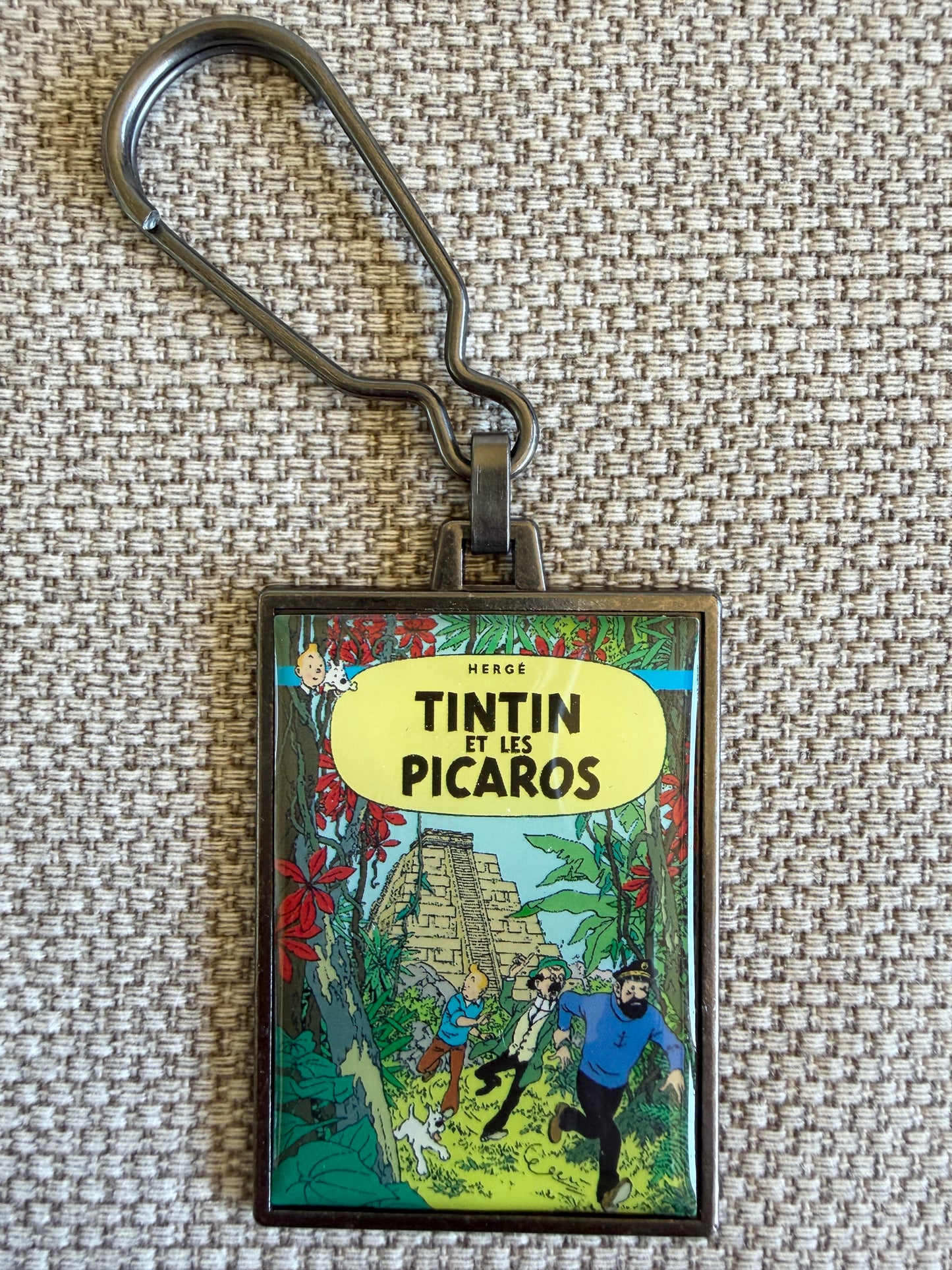 Front view of the rectangular Tintin Et Les Picaros metal keychain on a woven fabric background, featuring a resin-coated replica of the original French book cover showing Tintin, Captain Haddock, Professor Calculus, and Snowy running through a tropical jungle with an ancient Mayan pyramid in the background.