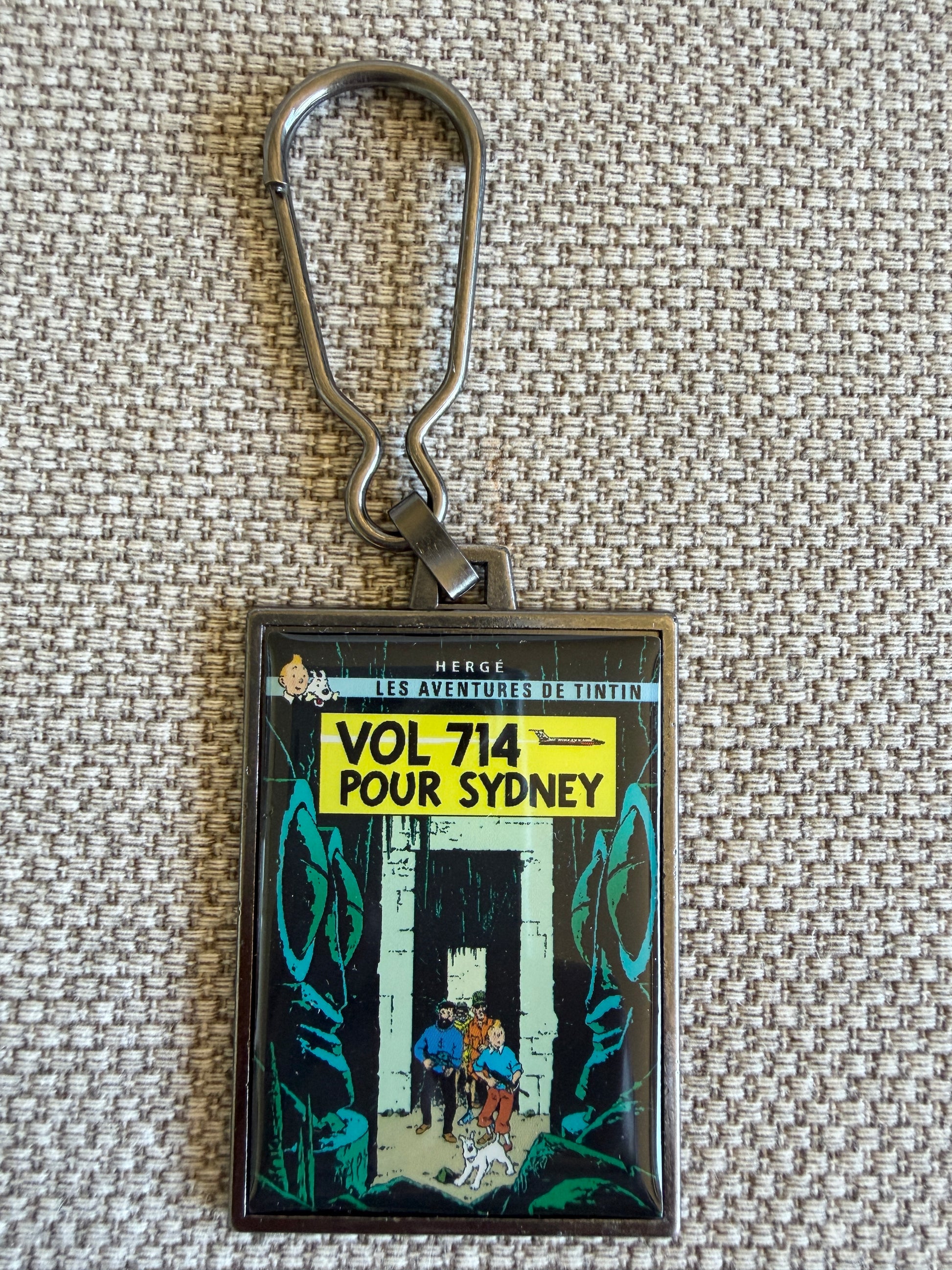 Front view of the rectangular Tintin Flight 714 to Sydney metal keychain, featuring a resin-coated replica of the original French book cover showing Tintin, Captain Haddock, and Snowy wading through a dense jungle.