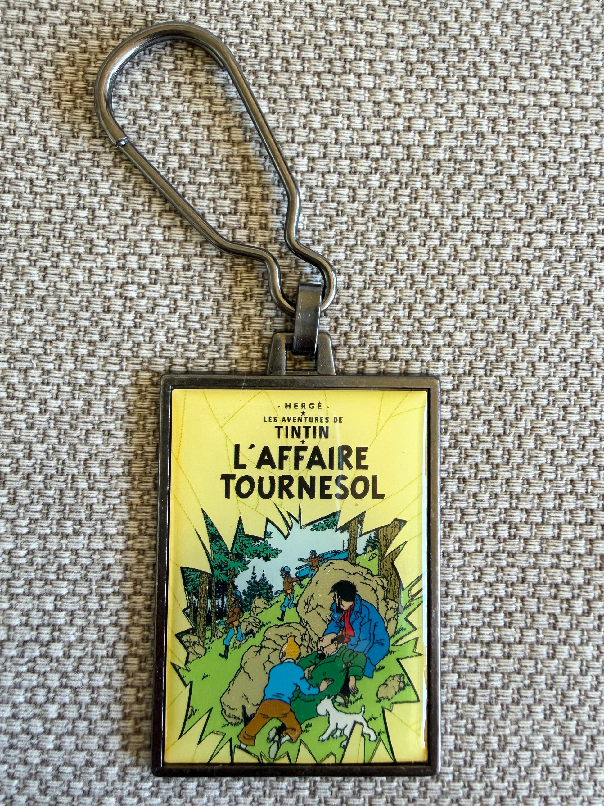 Front view of the rectangular Tintin L'Affaire Tournesol metal keychain on a textured, woven beige fabric background, featuring a resin-coated replica of the original French book cover showing Tintin, Captain Haddock, and Snowy crouching behind a large grey boulder while an armed border guard marches down a green hillside past them.