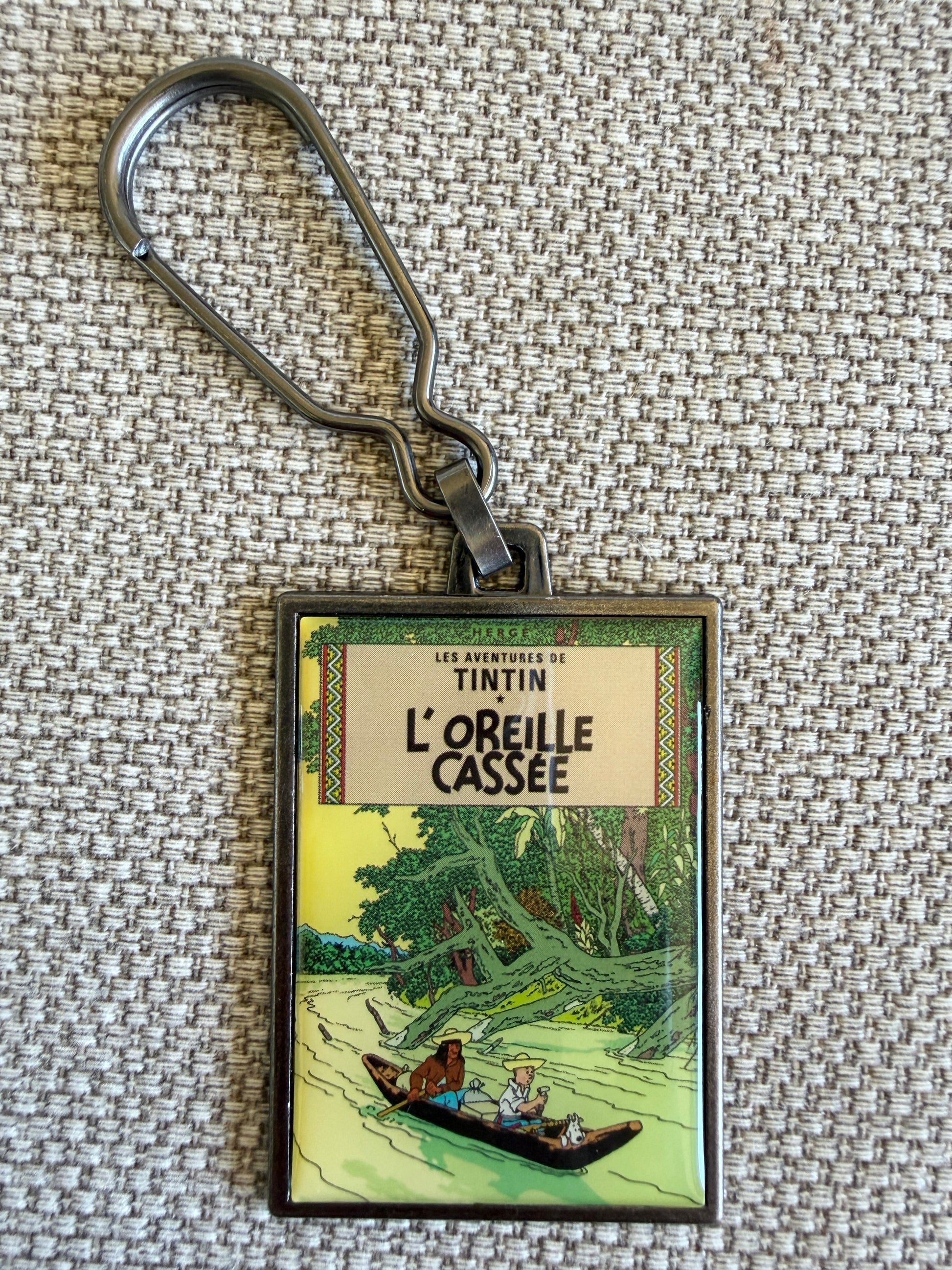 Front view of the rectangular Tintin L'Oreille Cassée metal keychain on a textured grey background, featuring a resin-coated replica of the original French book cover showing Tintin and Snowy navigating a tropical river in a wooden canoe surrounded by dense jungle foliage.