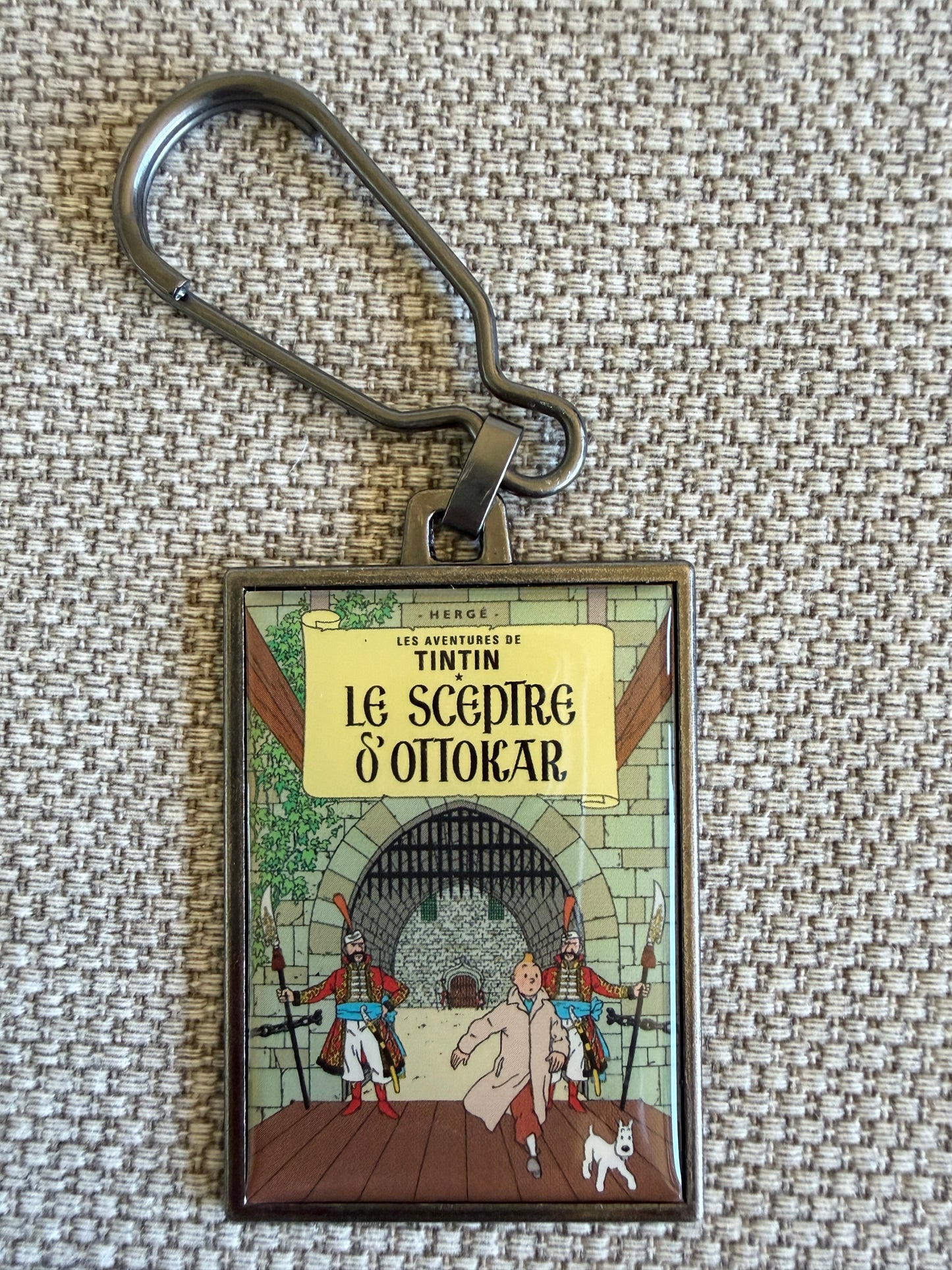 Front view of the rectangular Tintin Le Sceptre D'Ottokar metal keychain with a secure clip, featuring a resin-coated replica of the original French book cover showing Tintin running through a palace corridor with a Syldavian guard and Snowy.