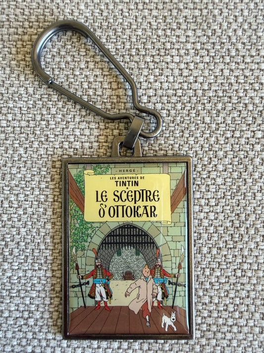 Front view of the rectangular Tintin Le Sceptre D'Ottokar metal keychain with a secure clip, featuring a resin-coated replica of the original French book cover showing Tintin running through a palace corridor with a Syldavian guard and Snowy.