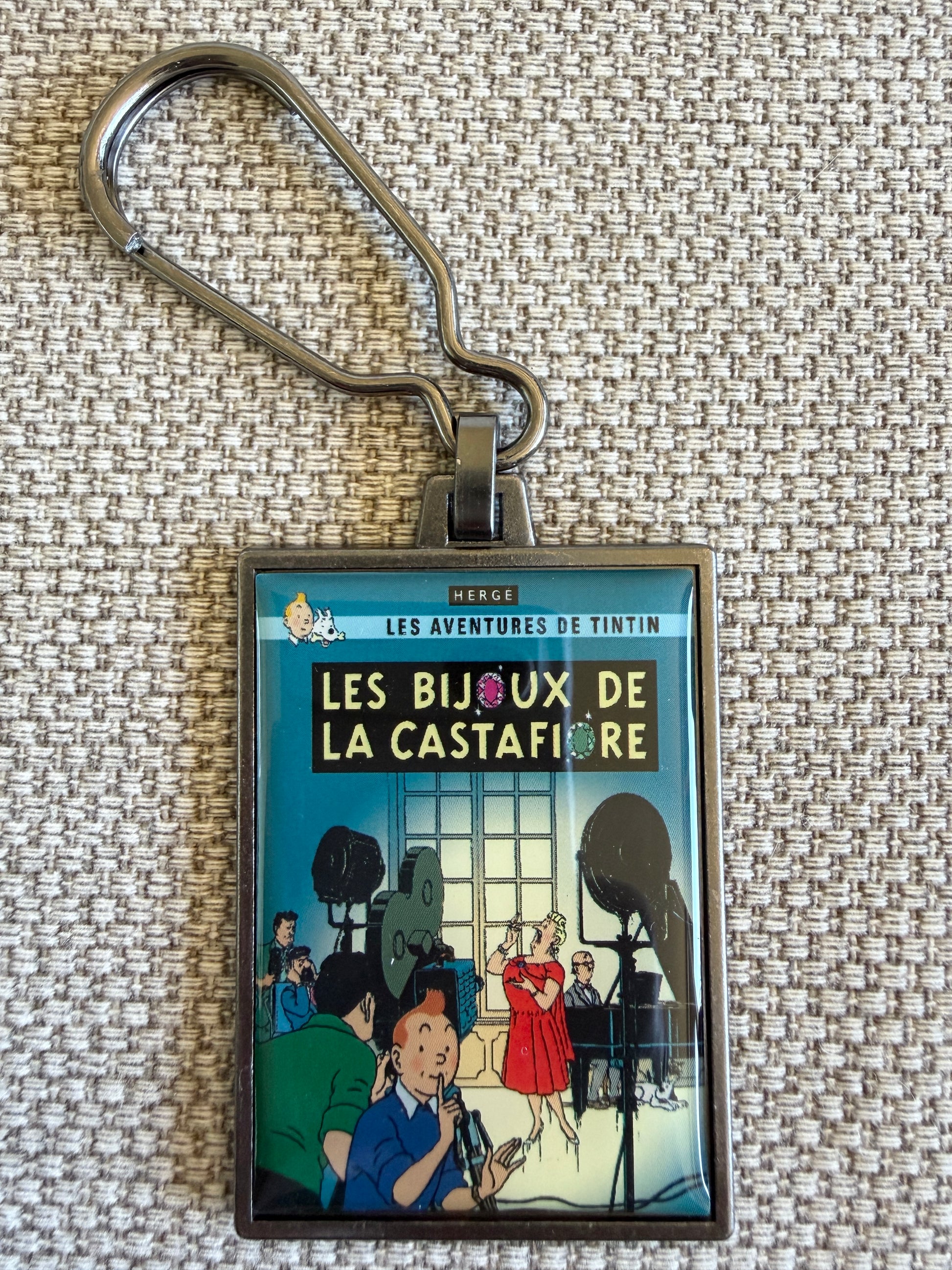 Front view of the rectangular Tintin Les Bijoux De La Castafiore metal keychain on a textured, woven beige fabric background, featuring a resin-coated replica of the original French book cover showing Tintin holding a finger to his lips to signal silence while a television crew films opera singer Bianca Castafiore in the background.