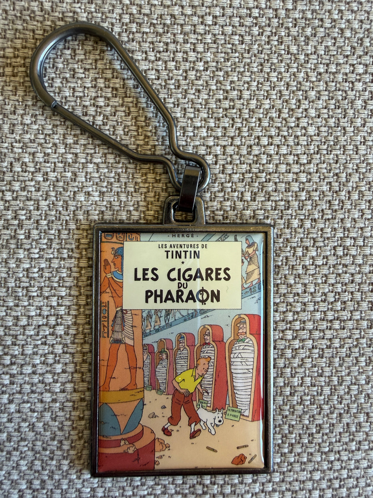 Front view of the rectangular Tintin Les Cigares Du Pharaon metal keychain on a textured beige fabric background, featuring a resin-coated replica of the original French book cover showing Tintin and Snowy walking down a stone corridor in an ancient Egyptian tomb lined with mummies in sarcophagi.