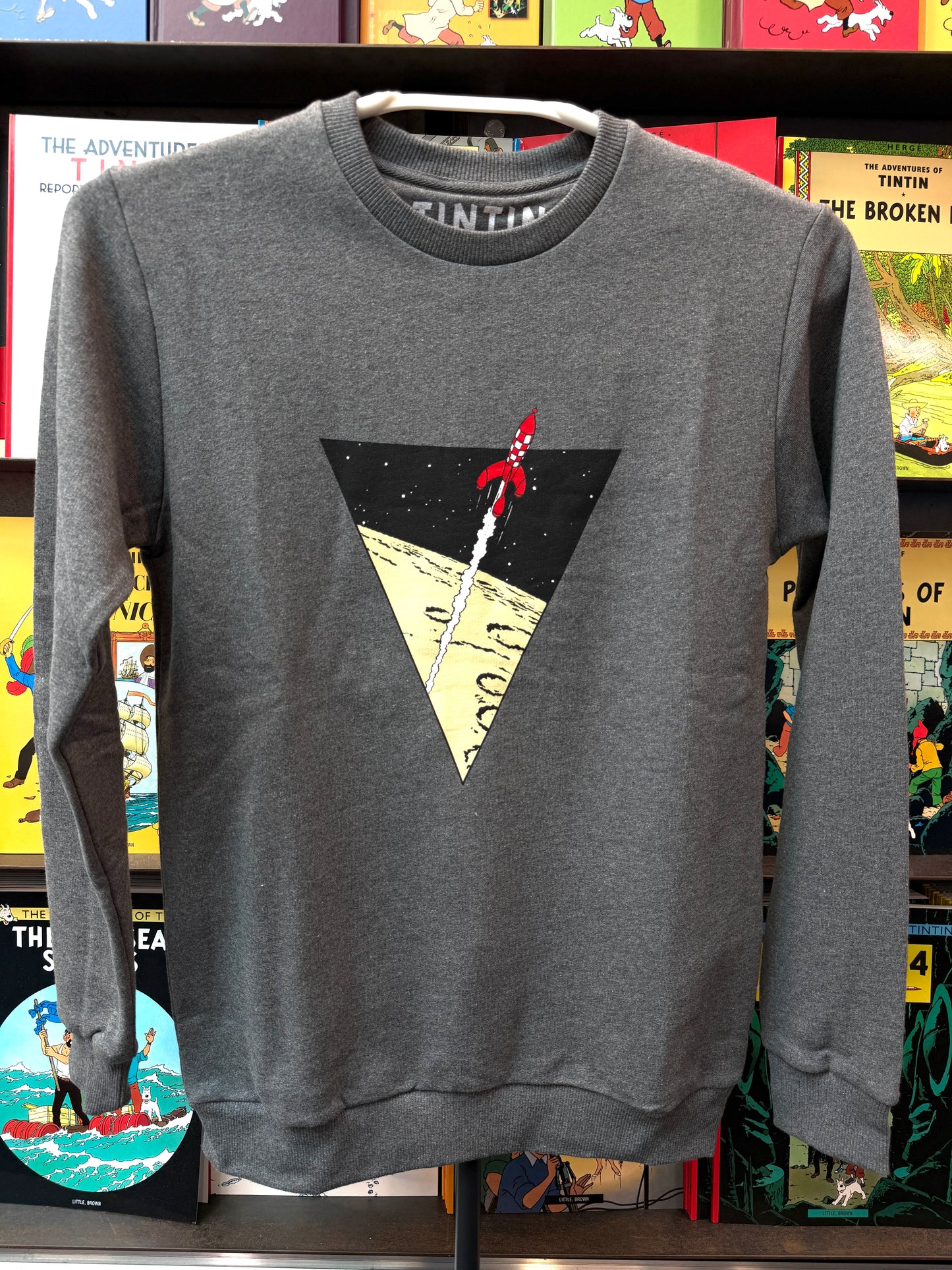 A dark heather grey unisex crewneck sweatshirt displayed against a colorful backdrop of Tintin comic books. The chest features a crisp graphic design of a large black inverted triangle framing the iconic red-and-white checkered moon rocket soaring upward, leaving a white smoke trail over a yellow lunar surface.