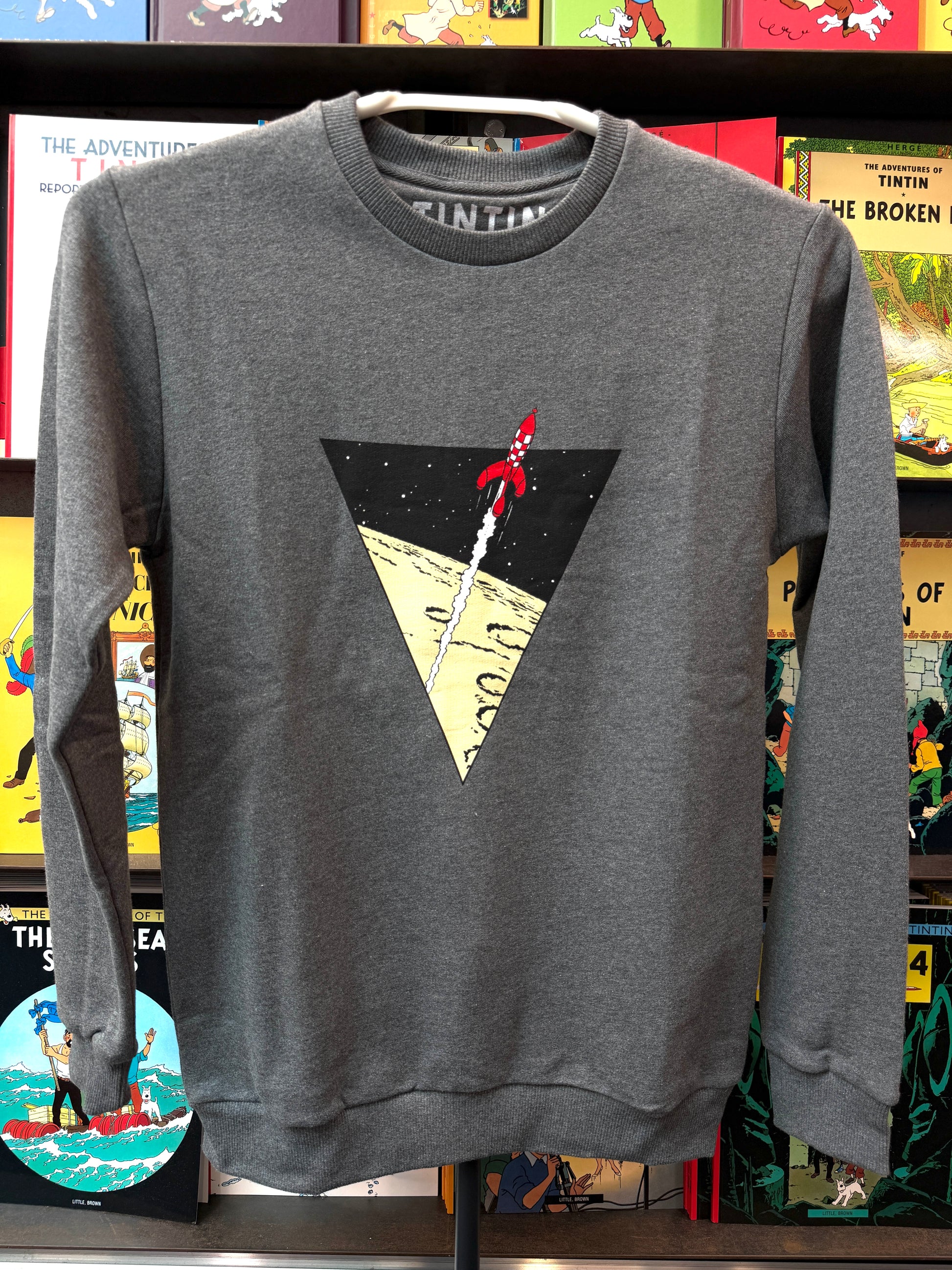 A dark heather grey unisex crewneck sweatshirt displayed against a colorful backdrop of Tintin comic books. The chest features a crisp graphic design of a large black inverted triangle framing the iconic red-and-white checkered moon rocket soaring upward, leaving a white smoke trail over a yellow lunar surface.