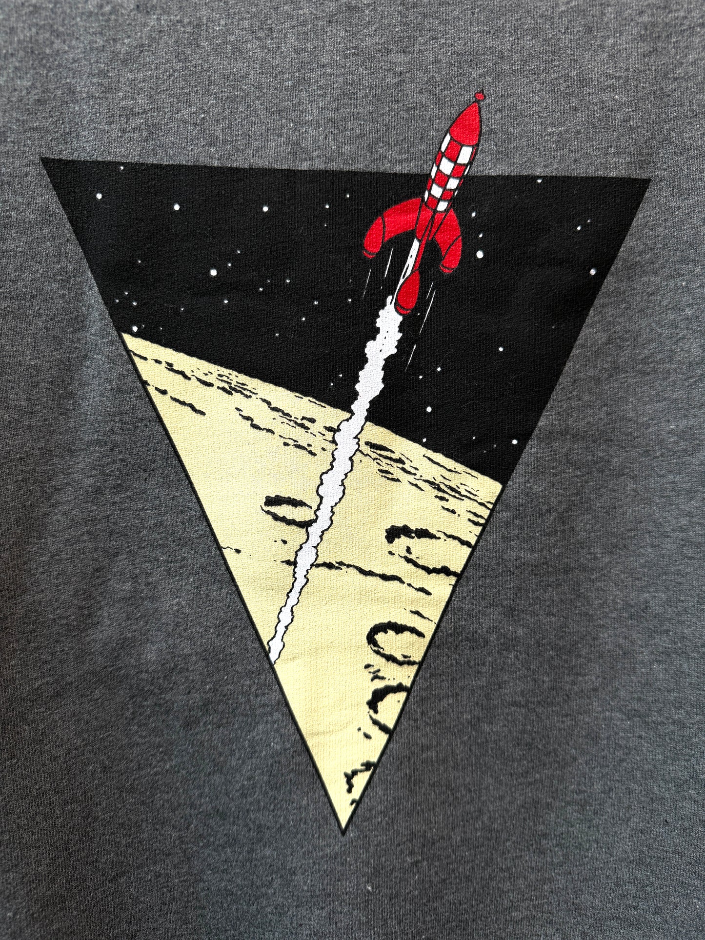 A close-up view of a dark heather grey textured fabric screen-printed with a large black inverted triangle. Inside the triangle, the iconic red-and-white checkered Tintin moon rocket launches upward into a black space filled with tiny white stars, leaving a long white trail of smoke over a light yellow, cratered lunar surface.