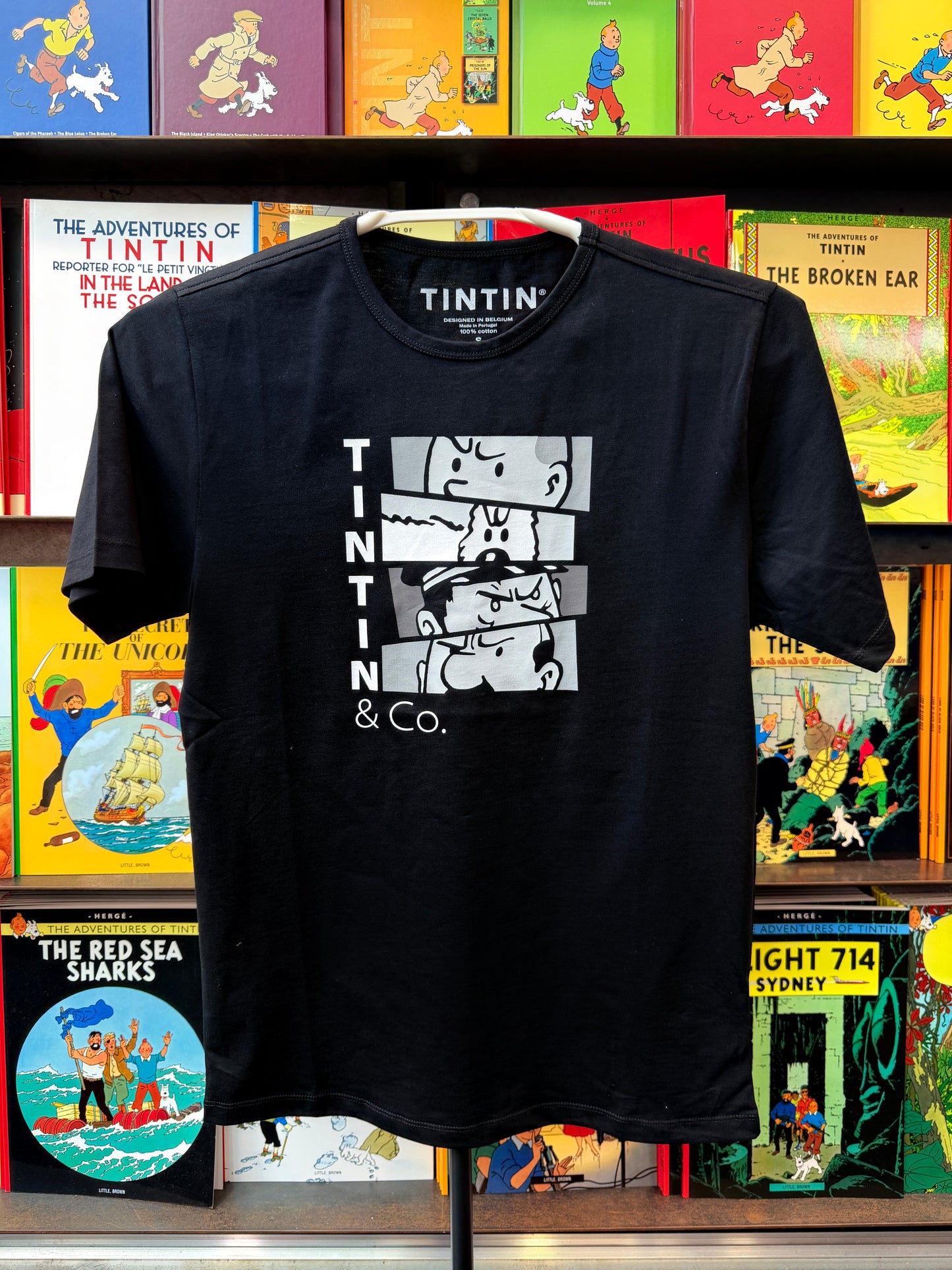 A front view of a black unisex t-shirt displayed against a background of colorful Tintin comic book spines. The t-shirt features a white graphic design on the chest composed of four stacked rectangular comic panels displaying close-ups of Tintin and Snowy, with the vertical text "TINTIN" stacked to the left and "& Co." printed cleanly underneath.