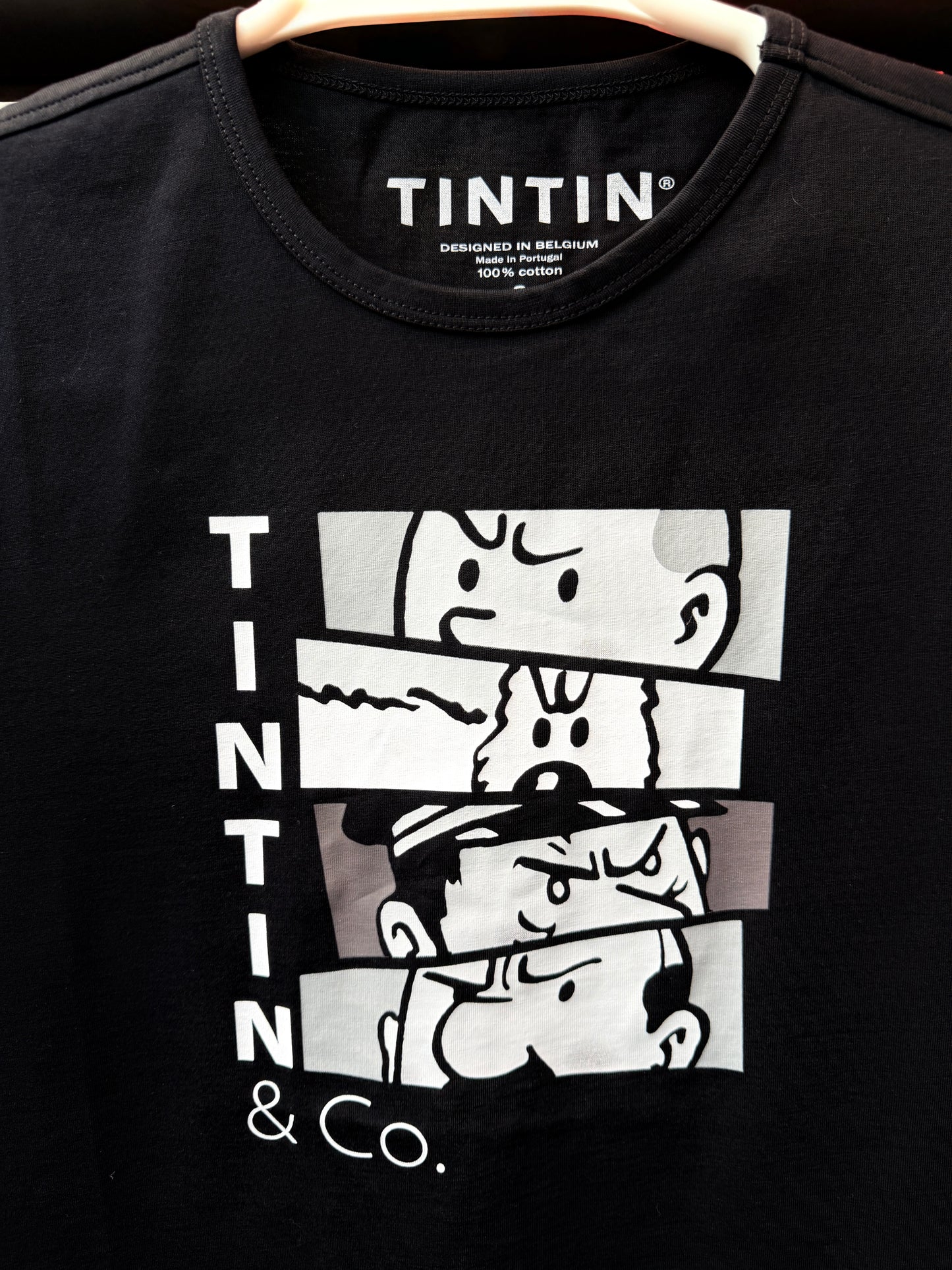 A close-up view of a black unisex t-shirt highlighting the white graphic print on the front. The design features the vertical text "TINTIN" next to four stacked horizontal comic panels showing dramatic close-ups of Captain Haddock, Snowy, and Tintin, with "& Co." printed cleanly underneath.