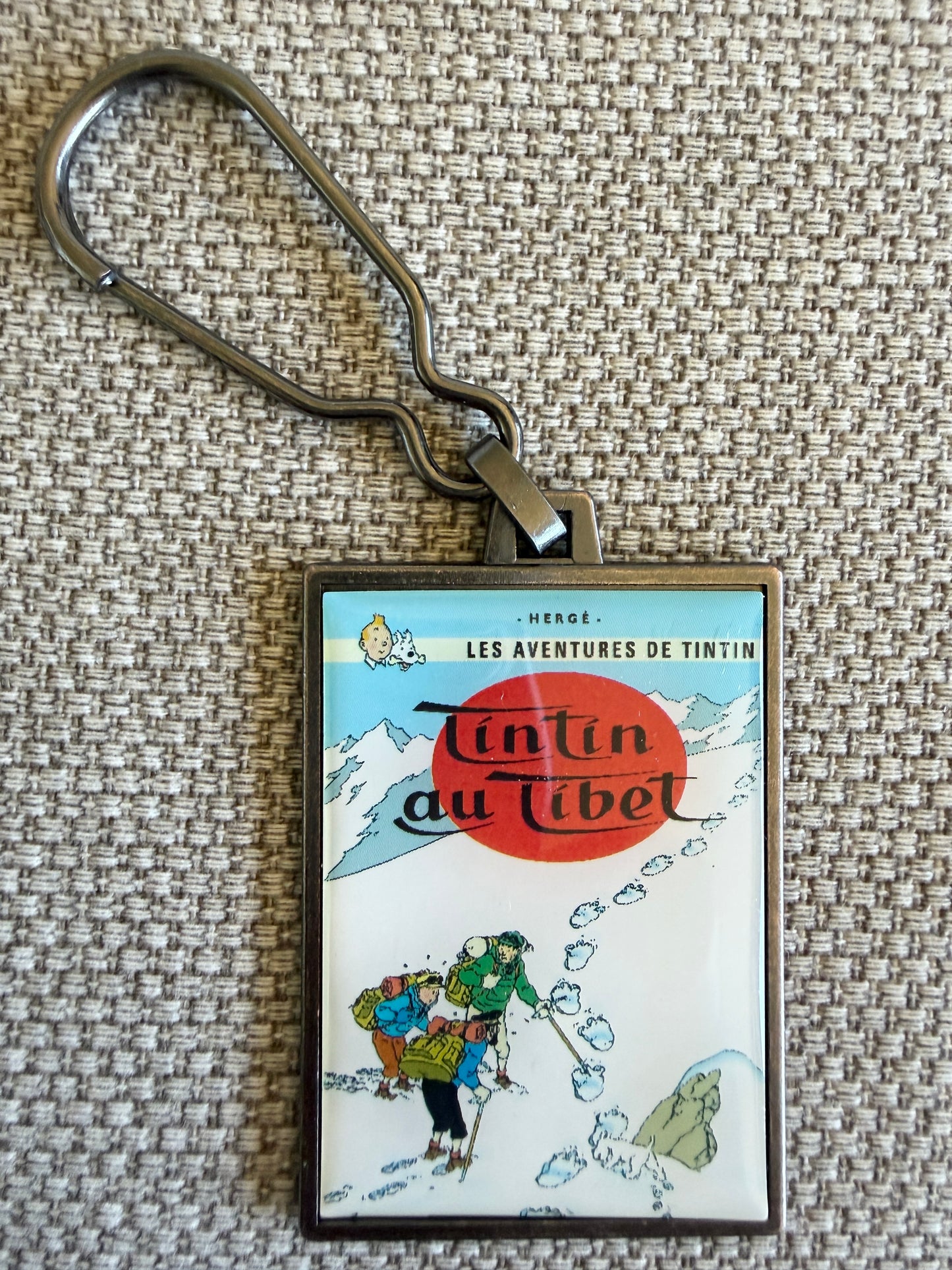 : Front view of the rectangular Tintin in Tibet metal keychain, featuring a resin-coated replica of the original French book cover showing Tintin, Captain Haddock, and Tharkey looking at a massive Yeti footprint in the snow.