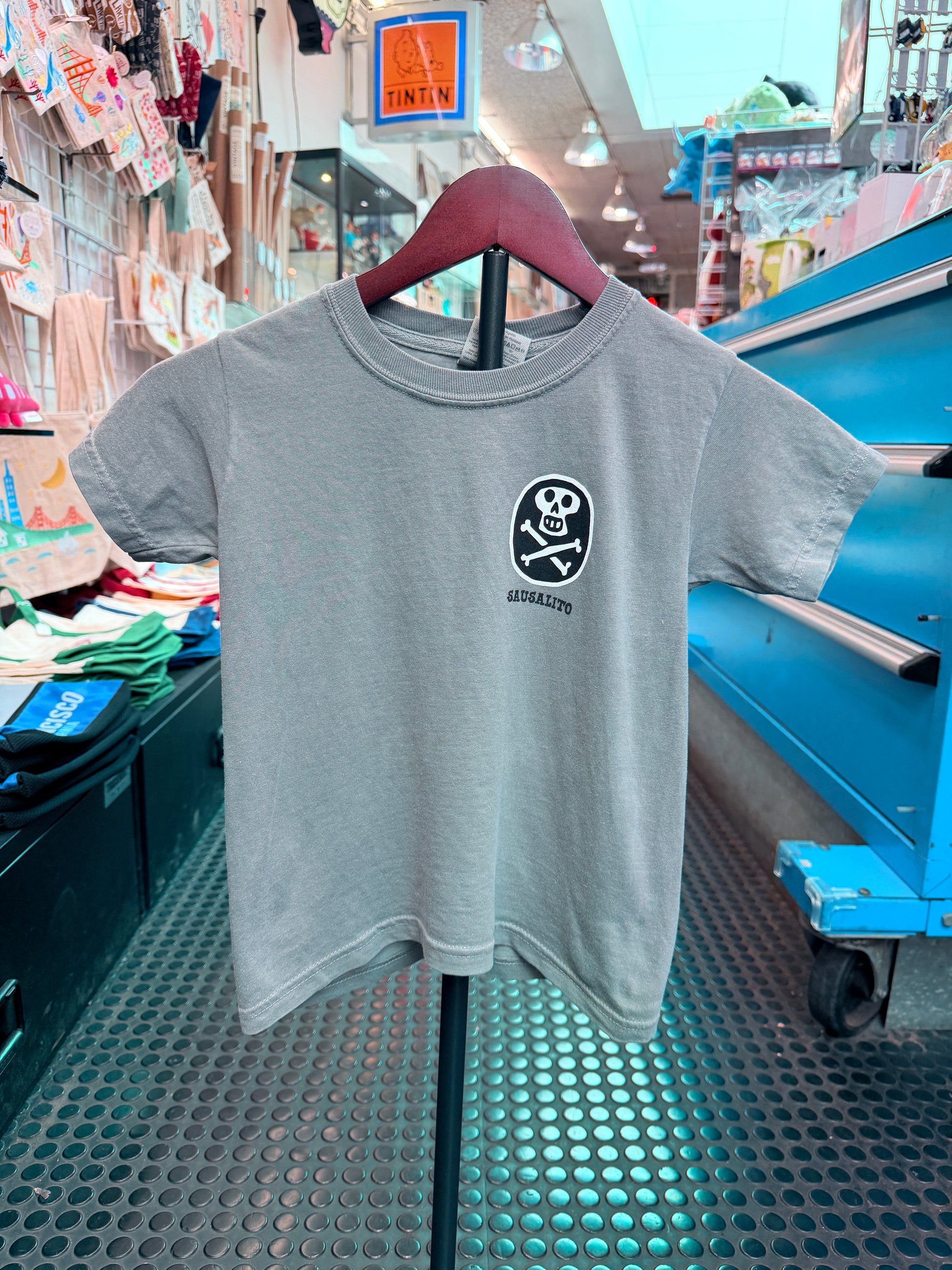 A light gray crewneck short-sleeve kid's t-shirt hanging on a display stand in a shop. The front chest features a small, black circular graphic containing a white skull and crossbones with the word 'SAUSALITO' printed directly underneath.
