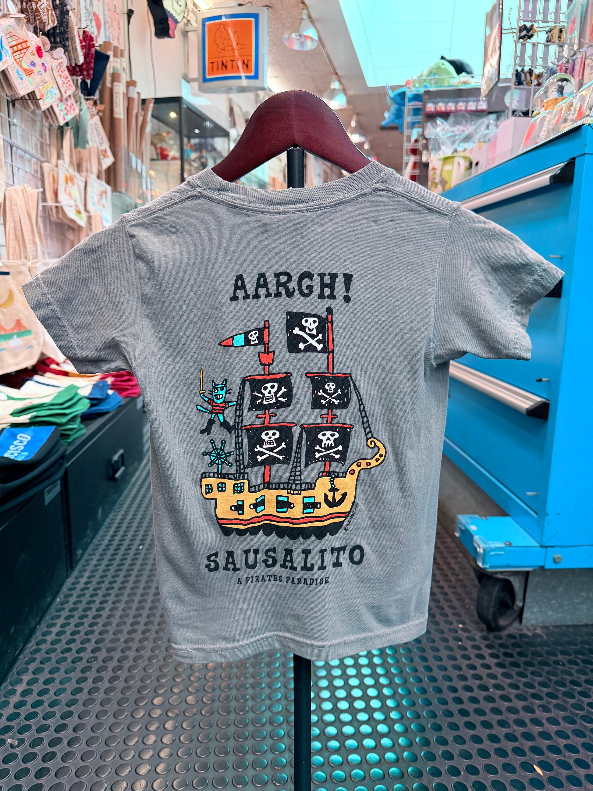 The back of a light gray short-sleeve kid's t-shirt hanging on a display hanger. It features a large colorful graphic of a whimsical pirate ship with black sails displaying white skulls and crossbones, a green monster holding a cutlass on deck, and bold text above and below reading 'AARGH!' and 'SAUSALITO A PIRATE'S PARADISE'.