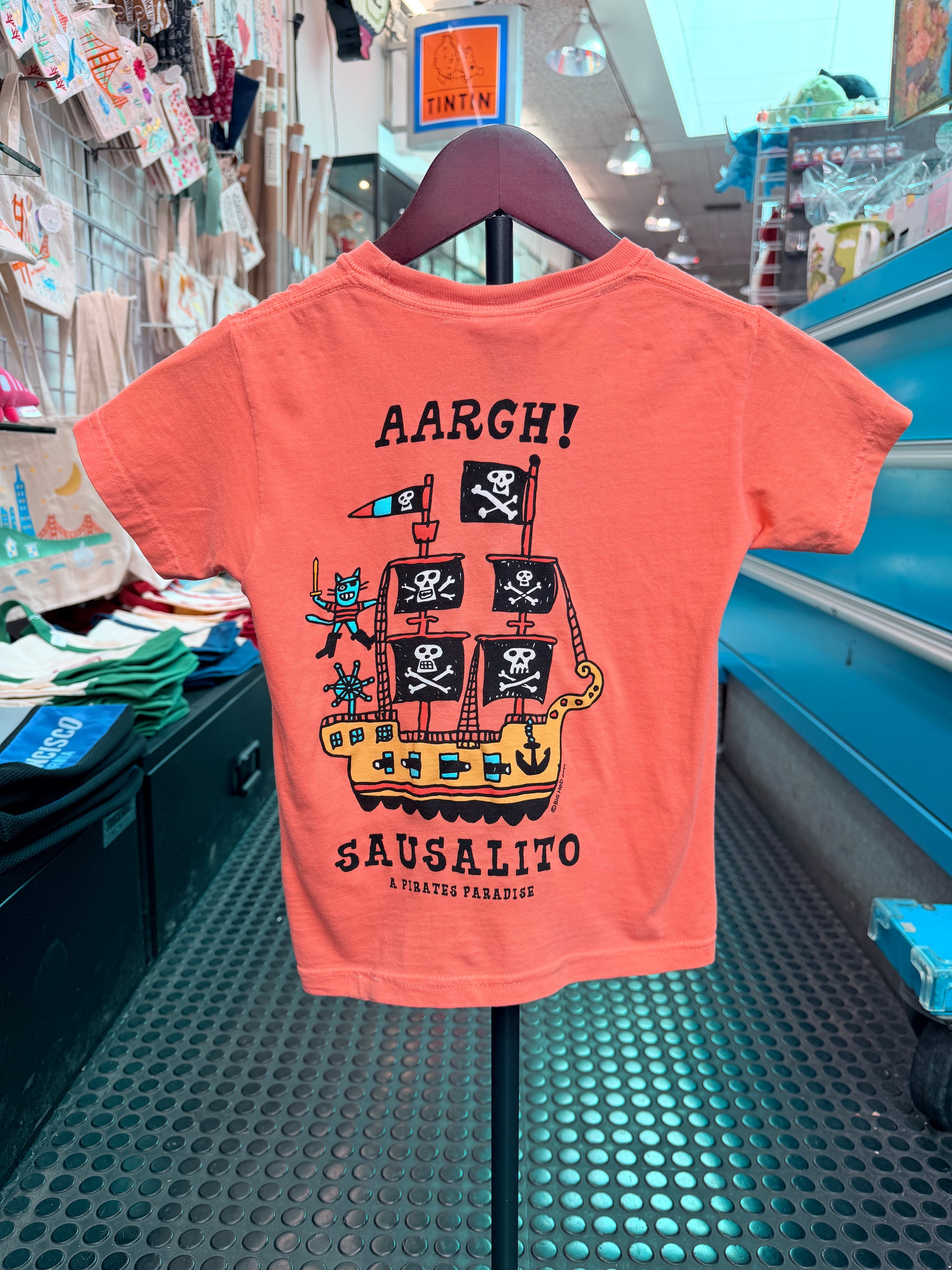 The back of a melon-orange short-sleeve kid's t-shirt hanging on a display hanger. It features a large black-and-white graphic of a whimsical pirate ship with skull-and-crossbones flags, a pirate cat holding a cutlass on deck, and bold text above and below reading 'AARGH!' and 'SAUSALITO A PIRATE'S PARADISE'.