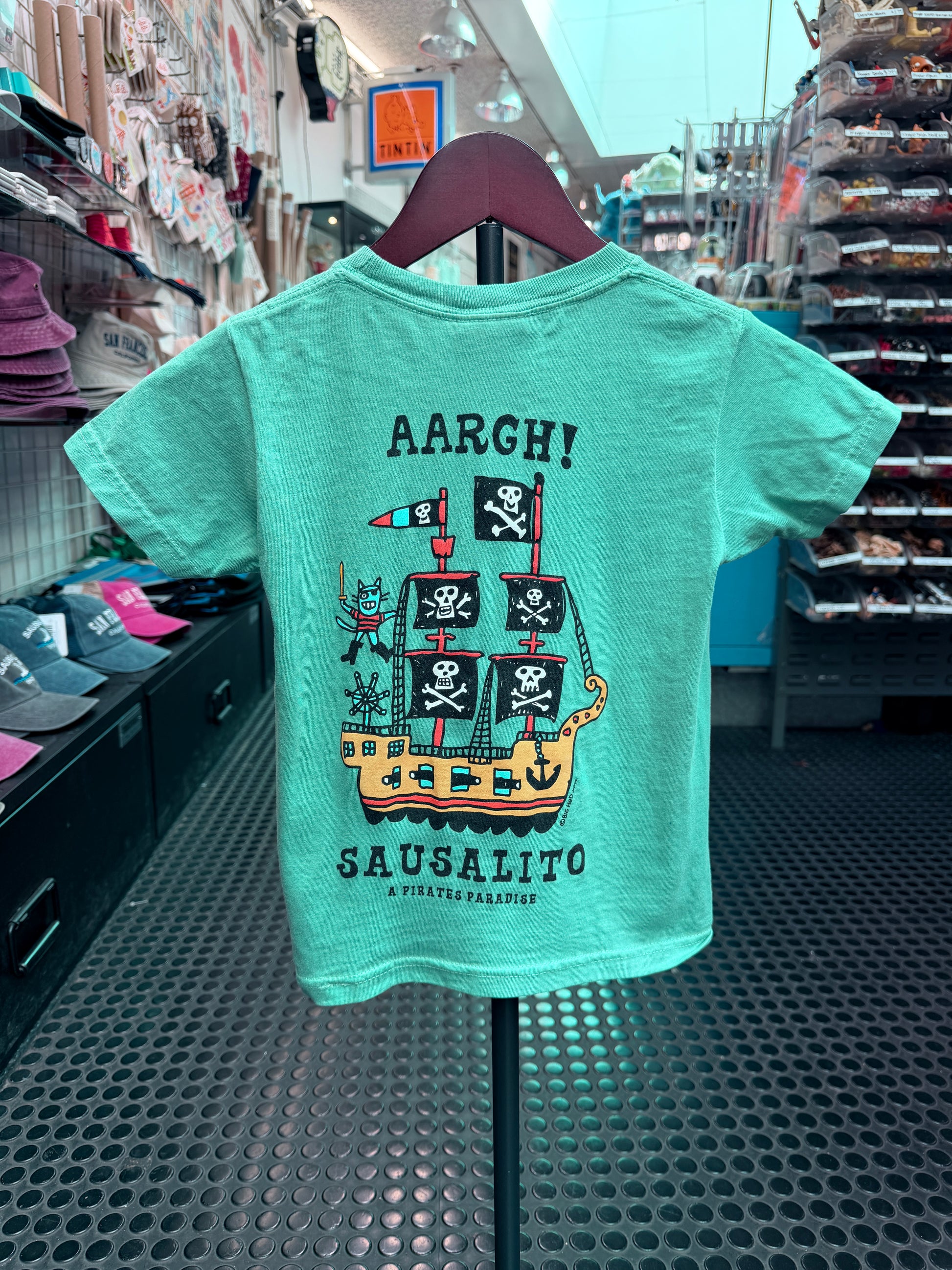 A mint green or seafoam short-sleeve kid's t-shirt hanging on a display, featuring a whimsical pirate ship graphic with skull-and-crossbones flags and the text 'AARGH! SAUSALITO'.