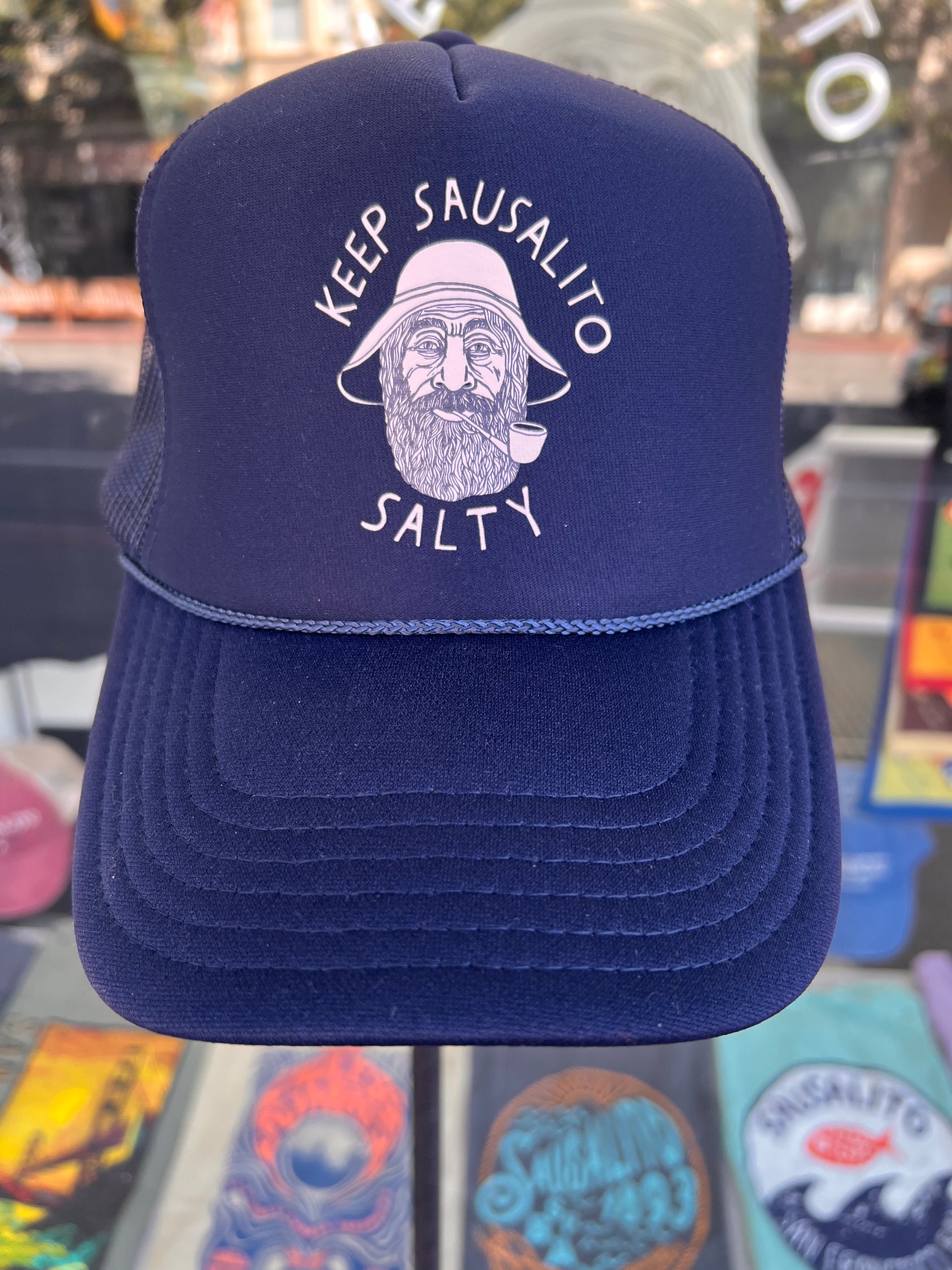 Keep Sausalito Salty Trucker Cap in navy blue featuring a white screen-printed graphic of a bearded fisherman in a rain hat with "Keep Sausalito Salty" text.