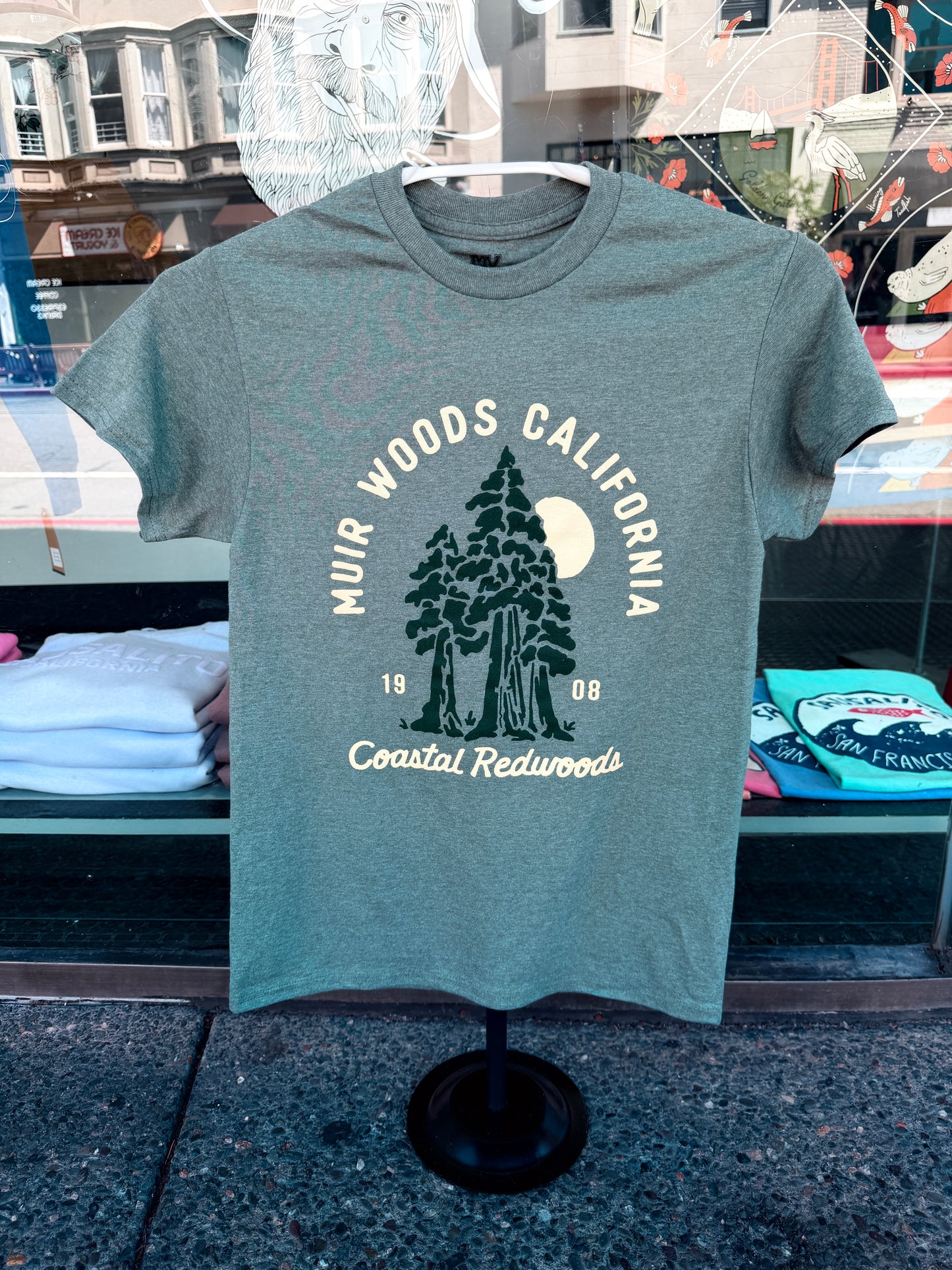 Muir Woods California Sunrise t-shirt in heather forest green displayed on a black clothing stand against a shop window reflection.