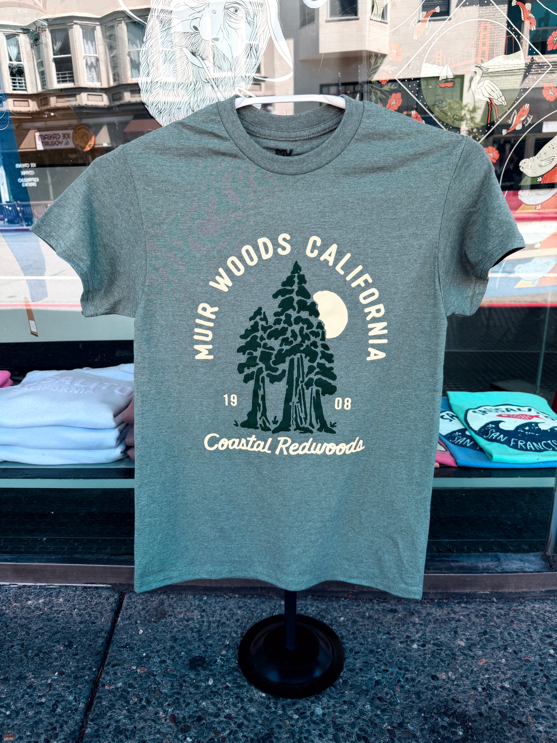 Muir Woods California Sunrise t-shirt in heather forest green displayed on a black clothing stand against a shop window reflection.