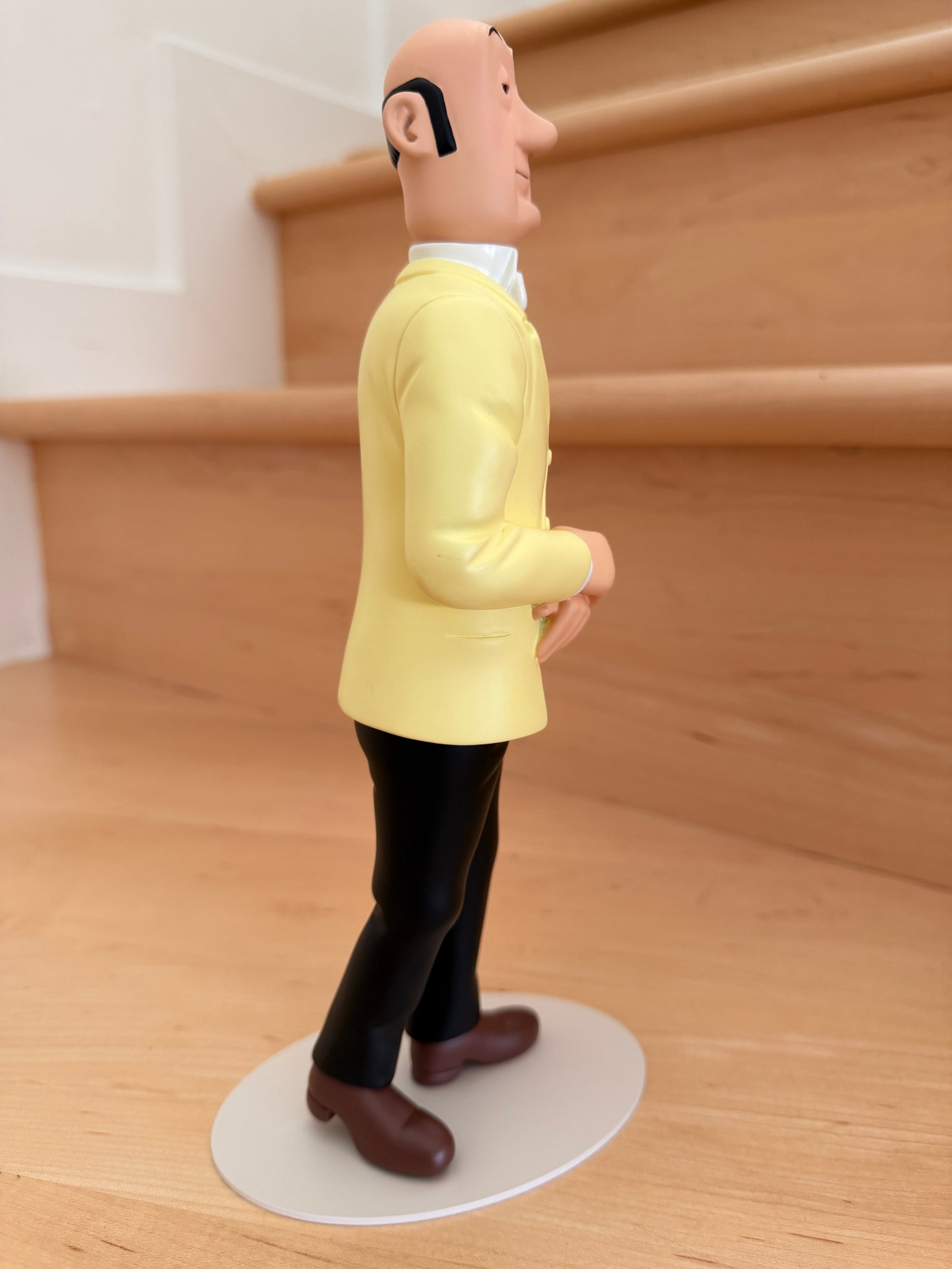 Profile view of the Tintinimaginatio Icons collection resin figurine of Nestor, the butler of Marlinspike Hall. He is depicted from the side with a calm, stoic expression, wearing his signature yellow double-breasted suit jacket and dark trousers, standing on a white circular base.